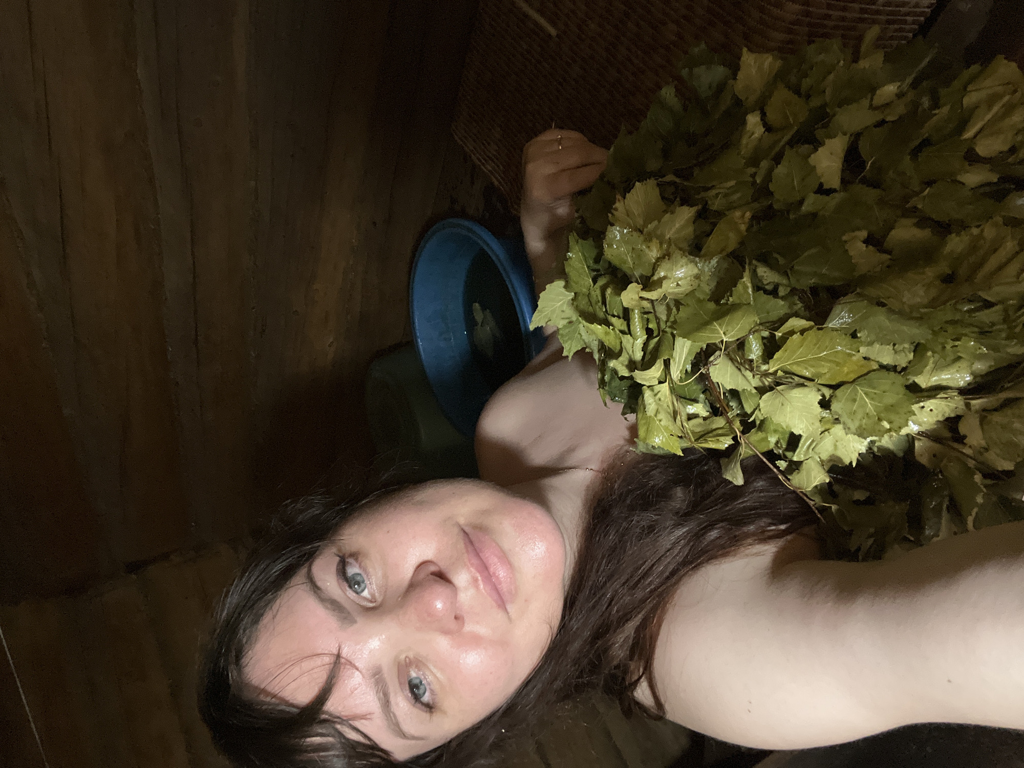 The image shows a woman in a sauna. She has brown hair and blue eyes and is holding a bundle of birch branches, which are commonly used in Russian banyas. The sauna appears to be constructed from wood. In the background, a blue bucket of water is visible, along with what appears to be a woven mat. The lighting is dim, creating a warm and intimate atmosphere. The woman is looking directly at the camera, with a slight smile.