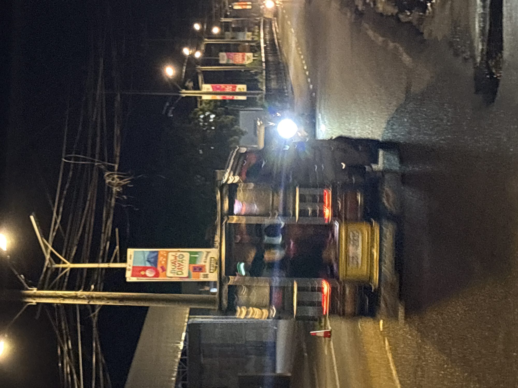 The image depicts a street scene at night in Davao City, Philippines, likely near a commercial or thoroughfare area given the presence of advertisements and street lighting. In the foreground, a blurred jeepney, a common mode of public transportation in the Philippines, is visible. It is dark with illuminated features, and passengers are visible inside, though their features are indistinct due to motion blur. The jeepney has a yellow rear bumper or chassis. Red taillights are illuminated. A traffic cone with reflective red and white stripes is placed on the road to the left of the jeepney.
In the background, a large pole with multiple wires is prominent, suggesting a complex urban electrical infrastructure. Attached to this pole are vertically oriented advertisements. One ad, more clearly visible, displays colorful graphics and text in red, yellow, and blue, with the words "DUAW. DAVAO." in bold lettering. Other similarly styled advertisements are also present, though less distinct. Streetlights illuminate the road and surrounding area with a warm, orange glow. A raised concrete barrier or sidewalk runs along the right side of the road. The road surface appears wet, reflecting the lights and creating a glistening effect. The overall atmosphere is nocturnal and urban, with a sense of movement and activity.