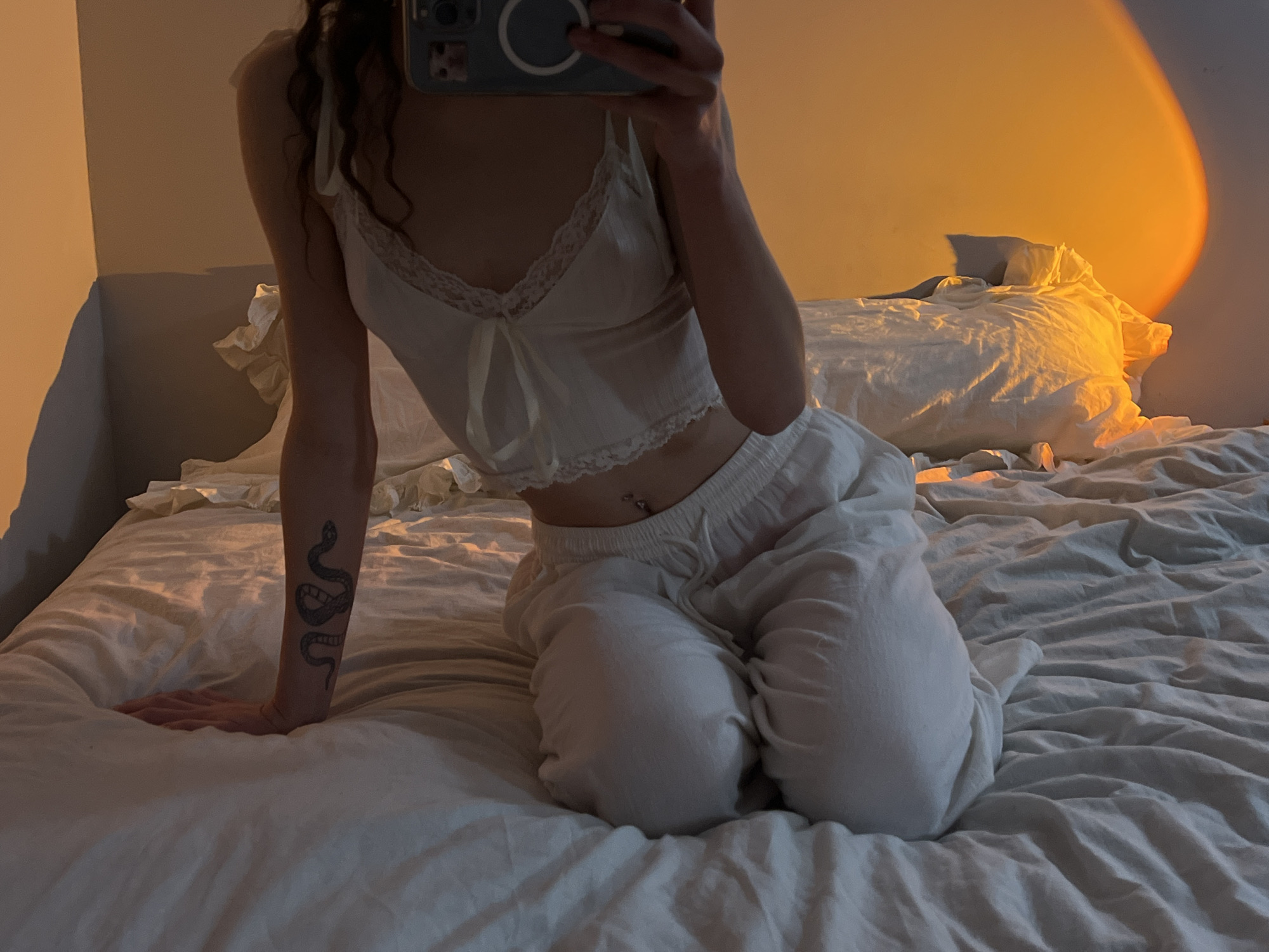 A person is kneeling on a white bed in a room illuminated by a golden light source that projects a halo on the wall. The person is wearing a white camisole with lace trim and white drawstring pants. They are holding a phone in their right hand, obscuring their face, and appear to be taking a photo. The person has a snake tattoo on their left forearm and a belly button piercing. The bed has white bedding and pillows. The location is indiscernible and cannot be identified as Ternopil, Ukraine based on the visual information provided.