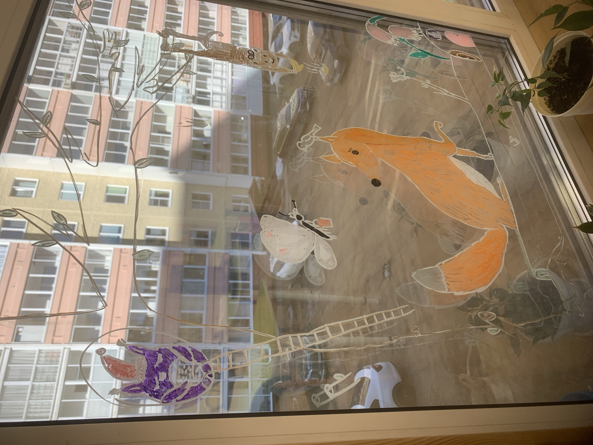 A child's artwork decorates a window with views of an apartment building, likely located in Izhevsk, Russia. The drawings on the window include an orange fox, a purple character inside a bubble, a butterfly, a ladder, and leafy branches. A potted plant sits on the windowsill. The reflection shows a multi-story apartment building with rows of windows. Cars are visible on the ground outside.