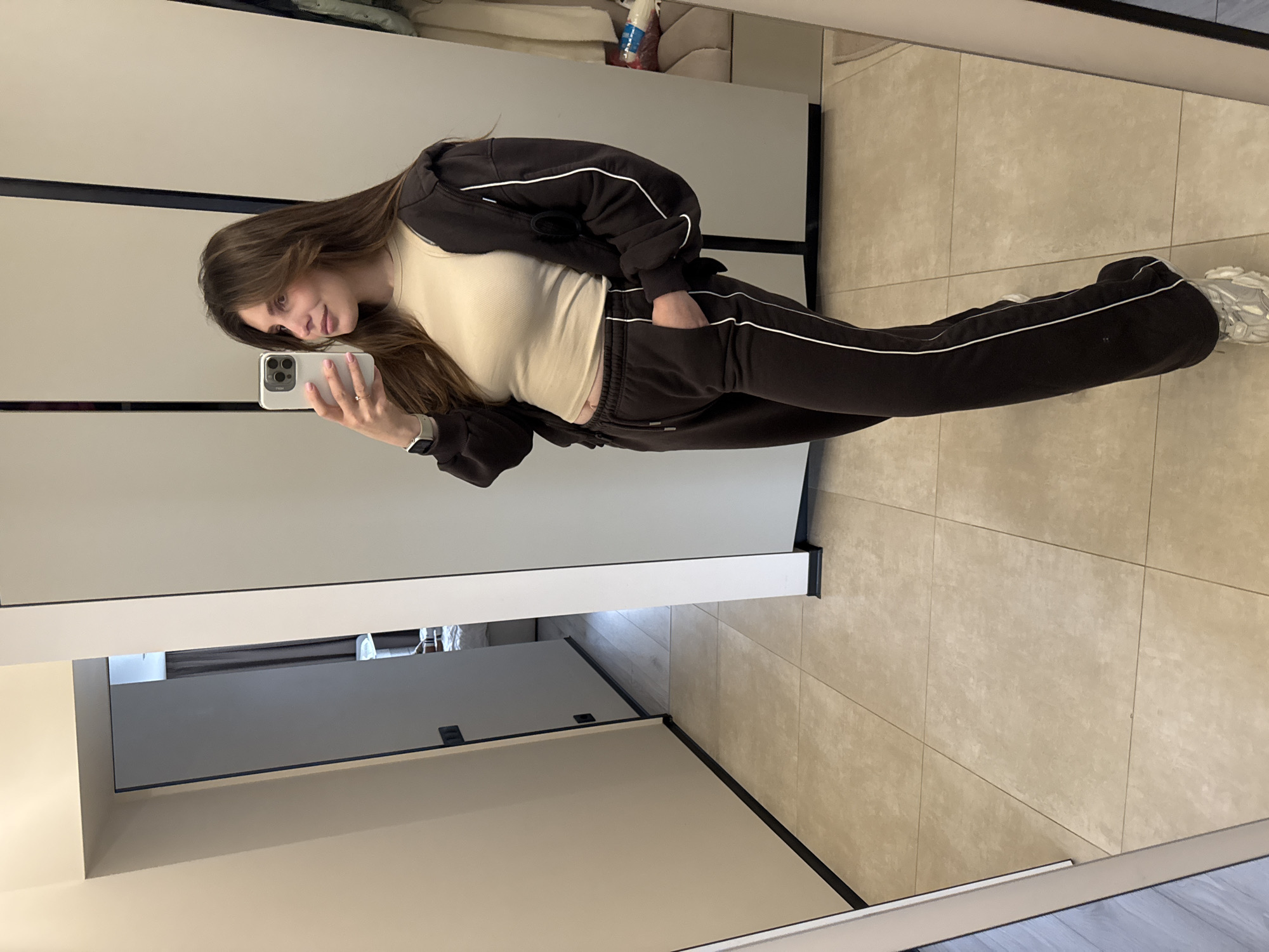 A young woman takes a mirror selfie in an indoor setting, likely in Sofiivska Borshchahivka, Ukraine. She has long, dark brown hair and is looking towards the phone with a subtle smile. She is dressed in a dark brown tracksuit with white stripe detailing along the sleeves and outer seams of the pants. Underneath her unzipped tracksuit jacket, she wears a light beige or off-white ribbed crop top, revealing a small portion of her midriff.
On her left wrist, she sports a white smartwatch, and her light-colored smartphone, possibly an iPhone, is held in her right hand, showcasing light pink painted nails. Her left hand is tucked into her tracksuit pocket. She completes her casual attire with chunky, light-colored athletic shoes.
The setting features a tiled floor with large, light beige or sand-colored rectangular tiles. The background consists of tall, neutral-toned panels or wardrobe doors, possibly light grey or off-white, reflecting the brightly lit interior. There are some indistinct objects visible in the reflection behind the camera's perspective, such as a white bottle. No discernible text is visible on clothing, the phone screen, or in the background. The overall impression is one of a relaxed, everyday moment captured in a well-lit indoor space.