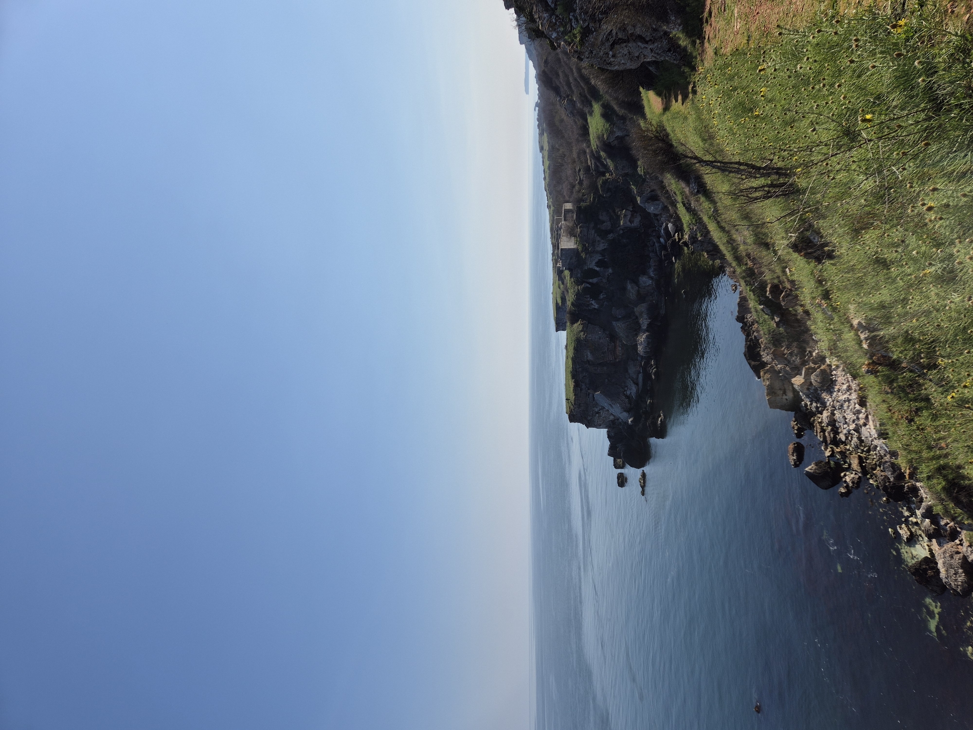 The image depicts a coastal landscape in Kamen Bryag, Bulgaria. The scene is split vertically: the left portion is a clear, blue sky. The right portion features a cliff face dropping into the sea. The cliff is topped with green grass and some sparse vegetation. Some rocky outcroppings extend into the water. The water appears calm and blue. There is a structure visible on the cliff, appearing to be ruins.