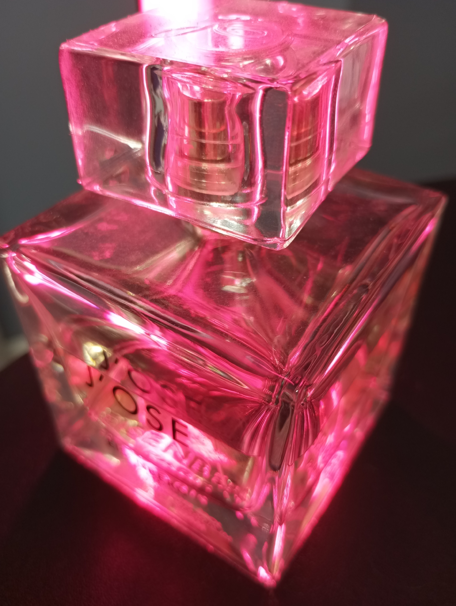 The image shows a close-up of a glass perfume bottle. The bottle is cube-shaped with a smaller cube-shaped cap. The bottle and its cap appear to be clear glass, though they are illuminated with a bright pink light, giving the impression that the glass is pink. A partial label reading "ROSE" is visible on the larger cube. The background is dark and out of focus, but there is a dark surface under the perfume bottle. The photo does not show the location in the city of Pervomaysky, Russia.