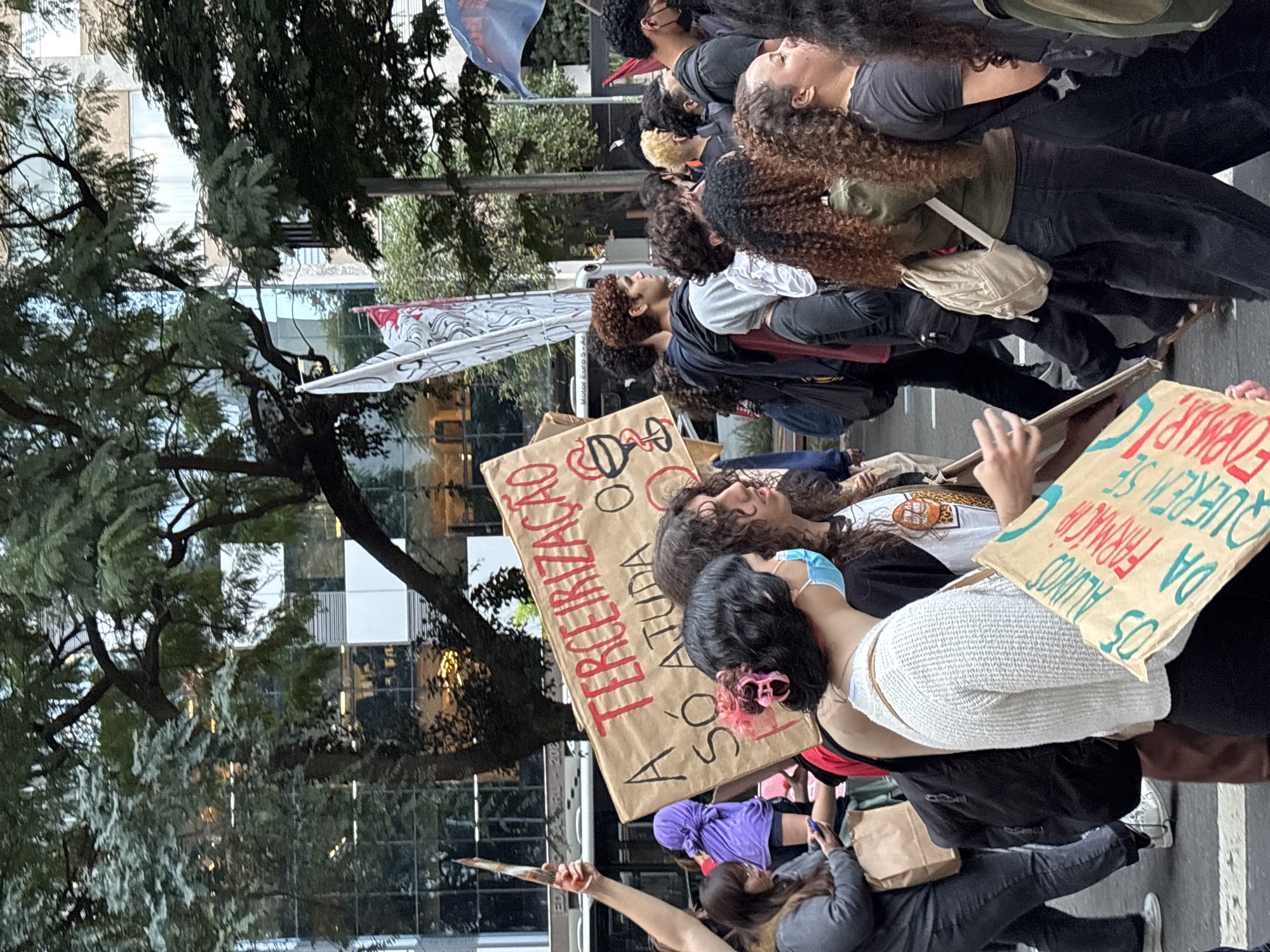 A group of people are marching, presumably a protest, on a street in São Paulo, Brazil. Several individuals are holding signs. One sign reads "A TERCEIRIZAÇÃO SÓ ATUDA", another says "05 ALUNOS DA FARMACIA QUEREM SE FORMAR". Trees and city buildings are visible in the background. Some individuals are wearing face masks. The group appears to be diverse. The setting is an urban environment during daylight hours.