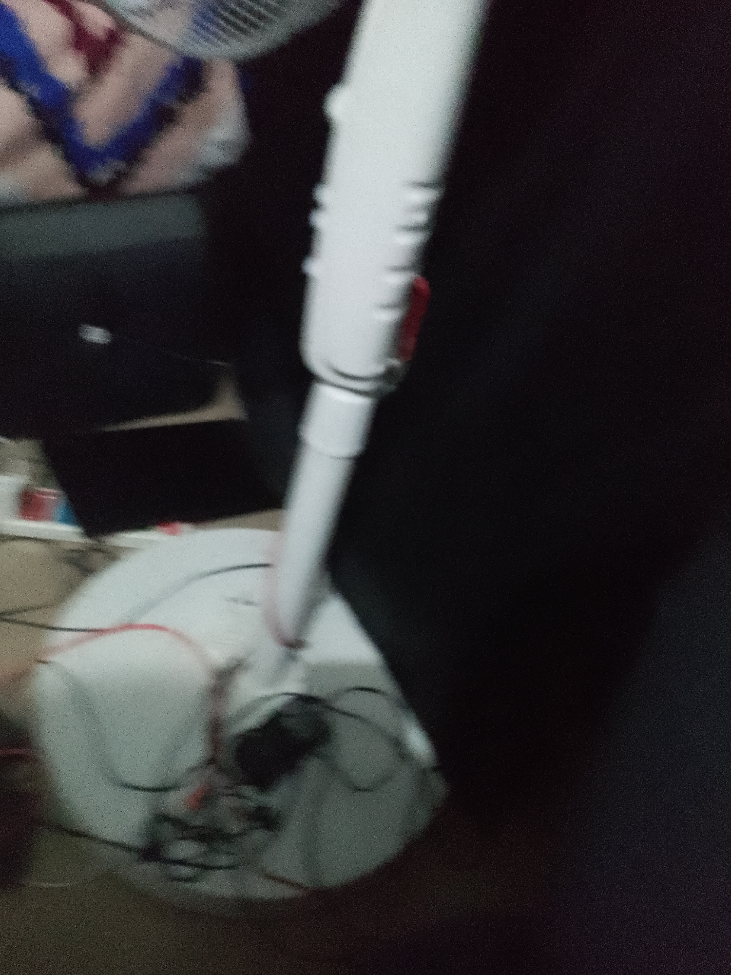 This is a blurry, low-light, close-up photograph of a white standing fan with its base visible at the bottom of the frame. The fan's pole extends upwards, and a red lever is visible on the side of the pole. The base of the fan appears to be made of white plastic and has some wires and a small black component attached to it. The background is dark and indistinct, with some blurry shapes that might be furniture or other objects. The image is highly pixelated and lacks clear detail. There is no identifiable location in Jalingo, Nigeria, or any discernible context beyond the fan itself.
