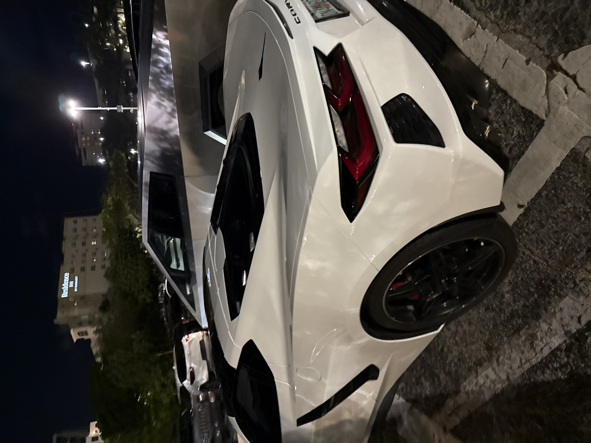 The image displays a white Chevrolet Corvette C8 sports car parked outdoors at night, viewed from a rear three-quarter angle. The car is sleek and modern, featuring prominent red LED taillights and black multi-spoke wheels with red brake calipers. A portion of the "CORVETTE" badging, reading "CORV", is visible on the rear deck lid. The vehicle is situated on dark asphalt with white parking lines.
The scene is set at nighttime, evidenced by the dark sky and the illumination from streetlights and building windows. In the background, several streetlights glow brightly, and various buildings are visible, with one prominently displaying the text "Residence Inn BY MARRIOTT". Trees line the distant landscape. The setting appears to be an urban or suburban parking area. No people are visible in the image.