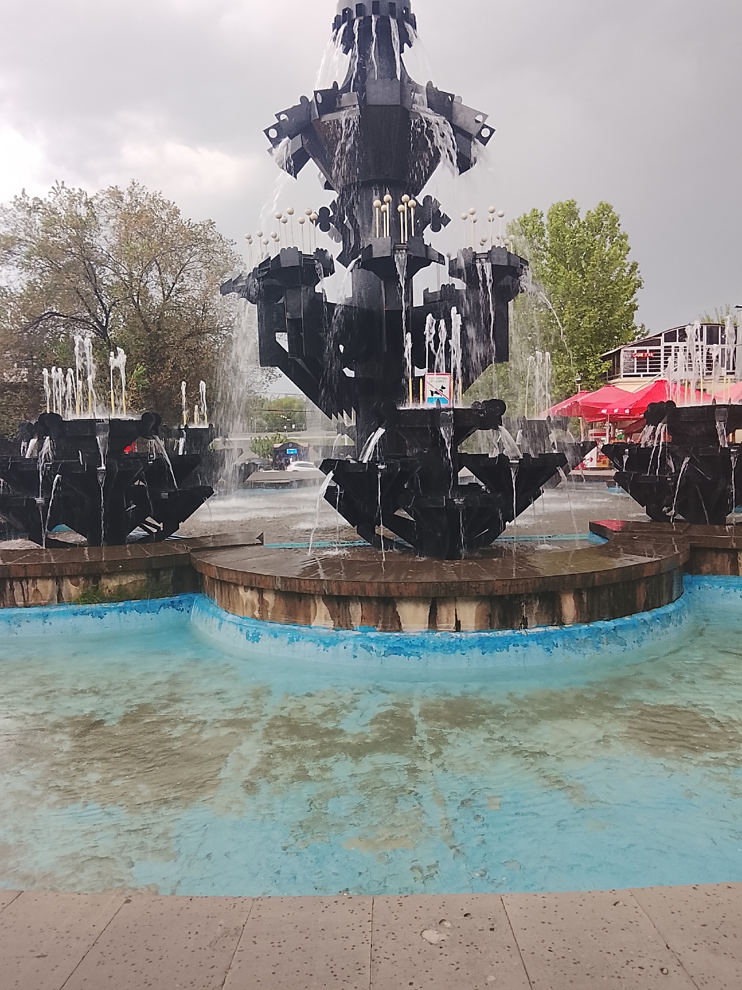 The image displays a large, elaborate multi-tiered fountain constructed from dark, geometric metal or stone, actively cascading water from various levels. The central structure features abstract, mechanical-like elements and small white spherical embellishments on thin rods. Surrounding the main fountain are several smaller, similarly styled dark fountain units, also emitting water jets. The fountain is situated within a pool with a light blue-painted interior lining, showing signs of wear and discoloration, particularly in the central water area which appears murky brown. The water level in the pool is relatively low, revealing parts of the bottom.
In the background, on the left, are deciduous trees with sparse foliage. On the right, more trees with greener leaves are visible. Behind the right side of the fountain, a building with large windows and a potential "GAME ZONE" sign is partially visible, along with a prominent red canopy or umbrella, suggesting a commercial or recreational area. The sky above is overcast and grey. The immediate foreground consists of a light grey, speckled paved surface, divided into rectangular slabs. No identifiable individuals are present. The location is the city of Unknown, Unknown.