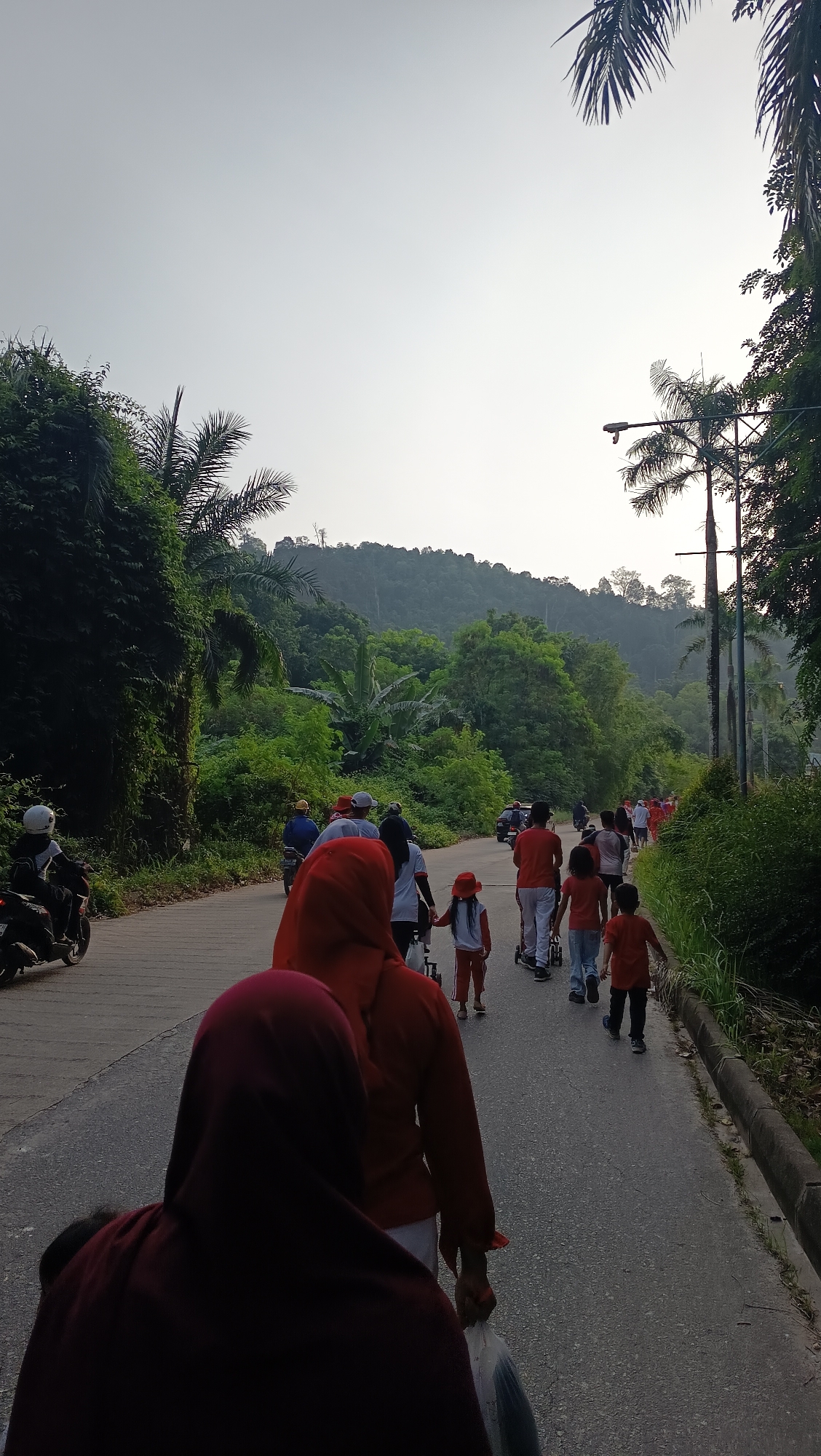 A group of people are walking on a paved road in a rural or park-like setting. The sky is overcast, and the lighting suggests it is either early morning or late afternoon. The road is lined with lush green vegetation, including trees and bushes. In the background, there is a hilly, forested area. Several individuals are visible, walking in the same direction on the road. Some are wearing red clothing. There are also a few motorcycles and a stroller present. The scene appears to be an outdoor recreational activity, possibly a walk or a communal event, set in a natural environment within or near Batam, Indonesia.