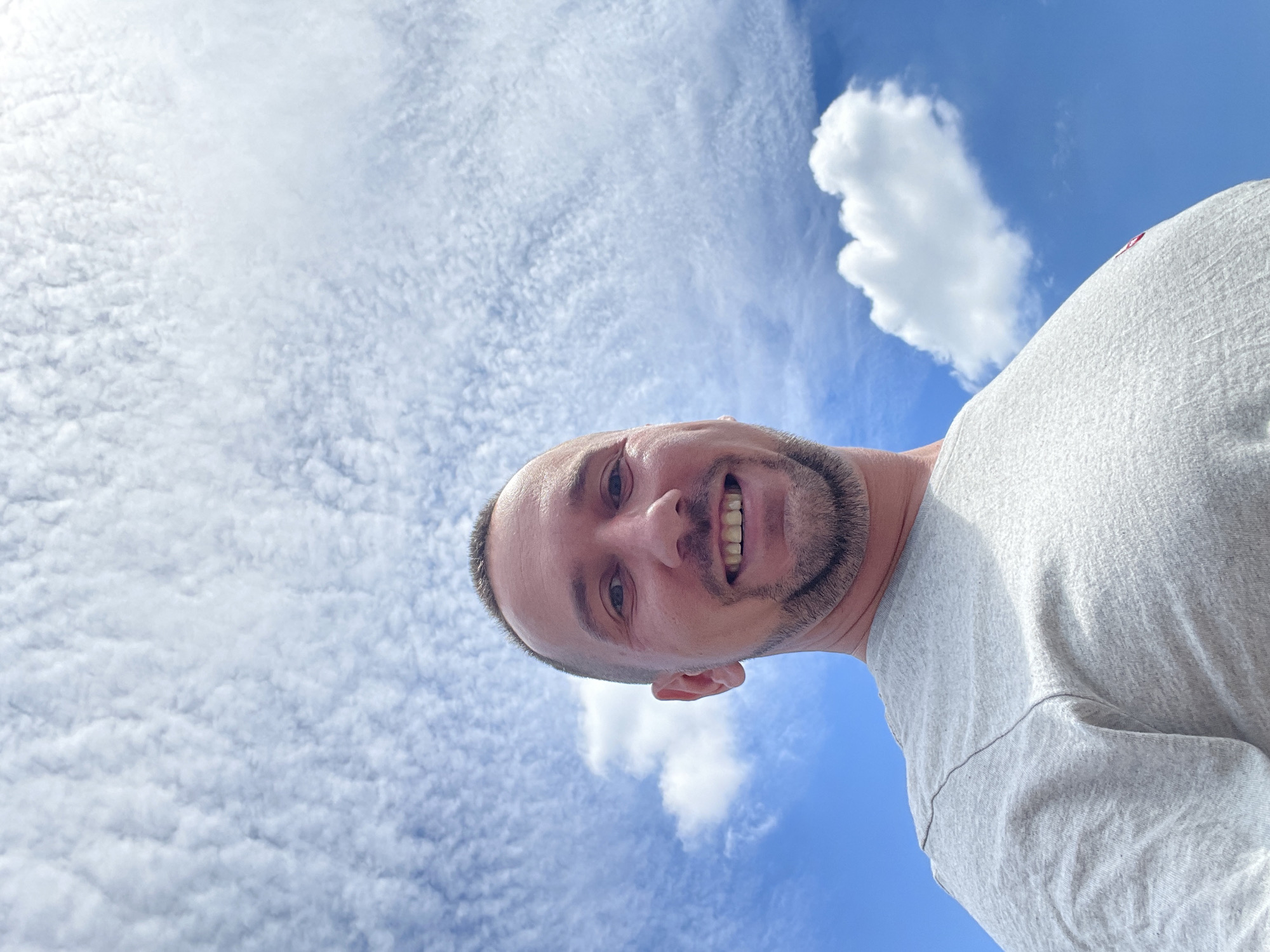 This is a low-angle, close-up, selfie-style photograph of a man against a backdrop of a bright blue sky filled with scattered white clouds. The man has short-cropped hair, a goatee, and is smiling. He is wearing a light gray t-shirt. The photo appears to have been taken outdoors, with the sky being the dominant feature of the background. There is no indication of the specific location within Kyiv, Ukraine.