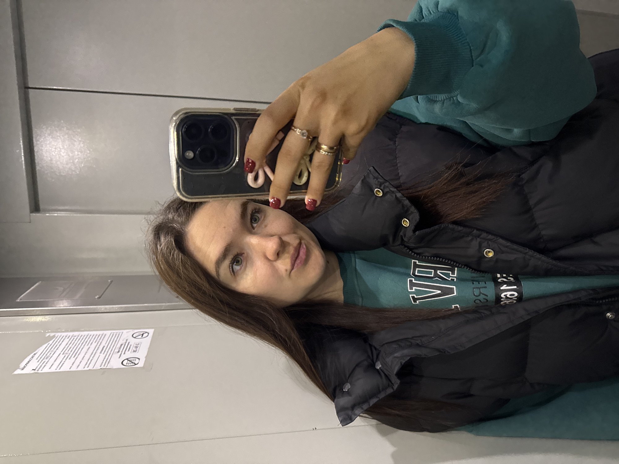 A young woman takes a mirror selfie in an indoor setting, likely an elevator or hallway, given the plain walls and small paper sign. She is looking directly into the phone camera with a calm, slightly smirking expression. Her long, dark brown hair frames her face, and her eyes appear light-colored, possibly green or hazel.
She holds a smartphone, likely an iPhone, with a clear case in her right hand, which displays several rings: a single one with a clear stone on her ring finger, and two simple gold bands stacked on her middle finger. Her fingernails are painted with glittery red polish. She is dressed in a dark teal or green long-sleeved top, possibly a sweatshirt, visible beneath a black puffer vest or jacket. The top has white, block letters, partially reversed in the mirror, reading "BA" and below it "ЯРЭ" (YARE) or similar Cyrillic letters, or possibly a distorted English word.
The background consists of light grey or off-white walls with horizontal seams, suggesting panels. In the lower left, a white paper sign with black text and symbols is affixed to the wall. Though mostly unreadable due to reflection and resolution, two circular symbols with diagonal lines are visible, one clearly indicating "no smoking" and another similar prohibition. The visible text on the sign appears to be in Cyrillic, consistent with the provided location context of Chaikovsky, Russia. The lighting is artificial and ambient, indicating an indoor environment.