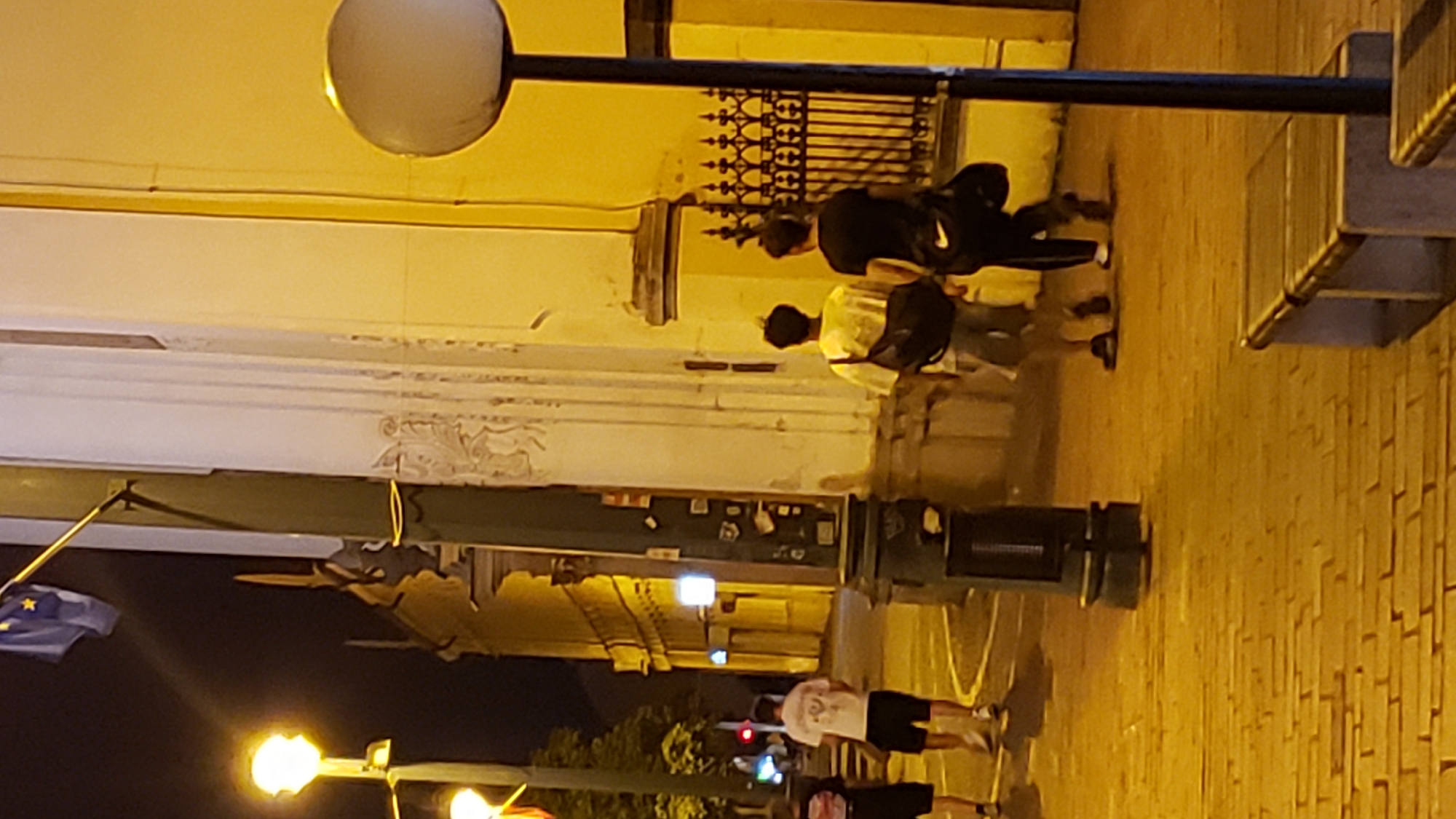 The image depicts a night scene on a cobblestone street, likely in the city of Prešov, Slovakia, given the context of the accompanying crops. The street is illuminated by streetlights and the warm glow from building interiors. In the foreground, two young men are walking away from the camera, one wearing a light-colored t-shirt and shorts, and the other a dark t-shirt and pants. Both are carrying bags. To the left, another person, partially visible, is also walking away from the camera. A lamppost with a round light fixture stands prominently on the right side of the image. Behind the pedestrians, a building with a yellow facade and ornate architectural details is visible, along with a decorative black metal fence. A European Union flag is also visible on a flagpole in the upper left portion of the image. The overall atmosphere is urban and active, suggesting a nighttime stroll through the city.