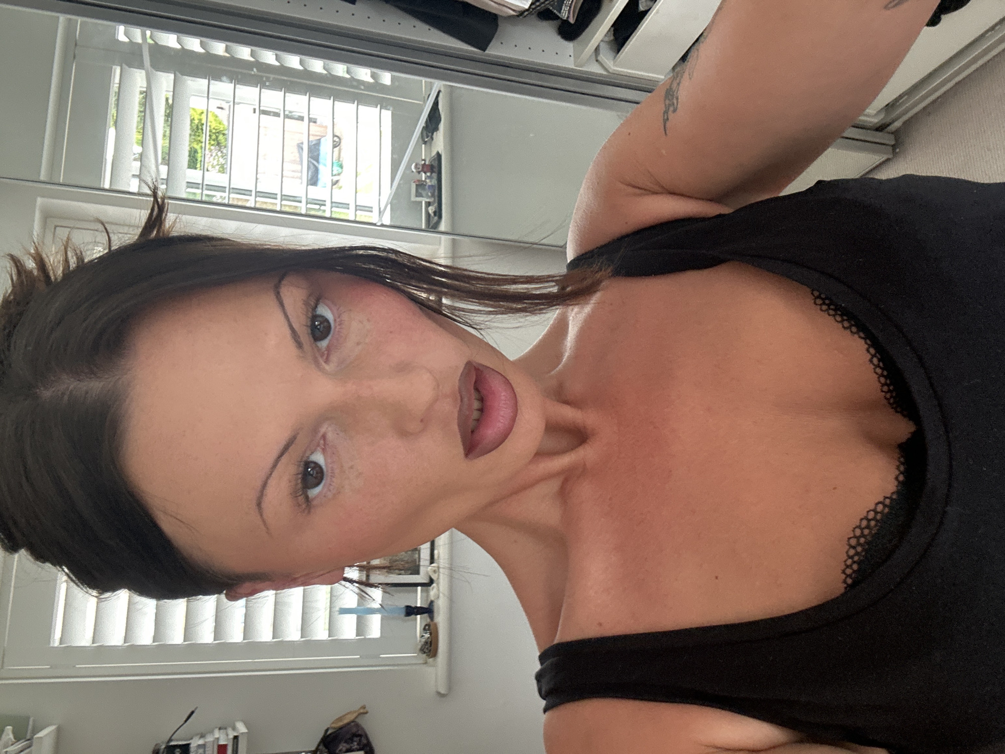 A woman is facing the camera, possibly taking a selfie. She has dark hair and is wearing a black top with lace detailing. The background appears to be a residential setting, with window blinds and what looks like a closet or wardrobe. The scene is indoors. The location in the city of Tunbridge Wells, United Kingdom, cannot be verified from the image.