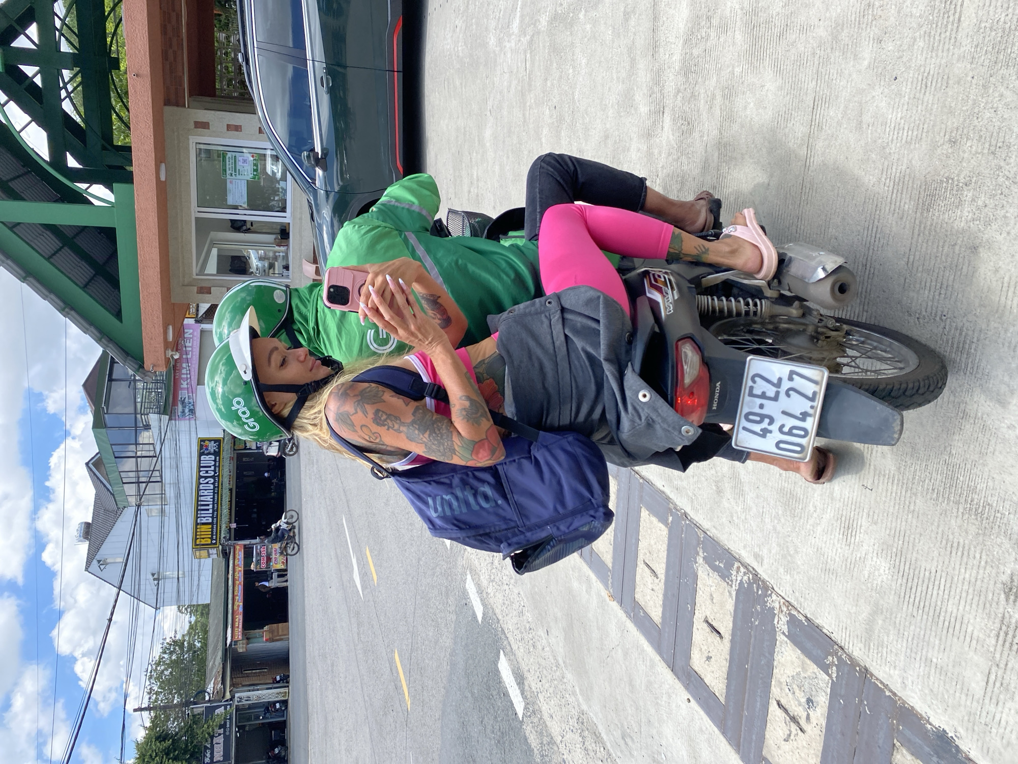 The image captures a street scene in Da Lat, Vietnam, on a cloudy day. In the foreground, a woman with blonde hair, wearing a pink and white top, black shorts, and pink leggings, is seated on the back of a motorbike. She has numerous tattoos on her arms and legs. She is holding a pink smartphone in her hands, seemingly taking a selfie or checking her device. She is wearing a green helmet with the "Grab" logo visible. The motorbike is black and gray with a visible license plate that reads "49-E2 064.27". Behind her, a person wearing a green Grab jacket and a green helmet is driving the motorbike. The driver is wearing dark pants.
The background features a street with white lane markings. To the left, there are buildings with signs, including one that reads "BIII.M BILLIARDS CLUB" in yellow lettering. A dark green SUV is parked to the right. Overhead, power lines crisscross the sky. The overall environment suggests an urban setting within Da Lat.