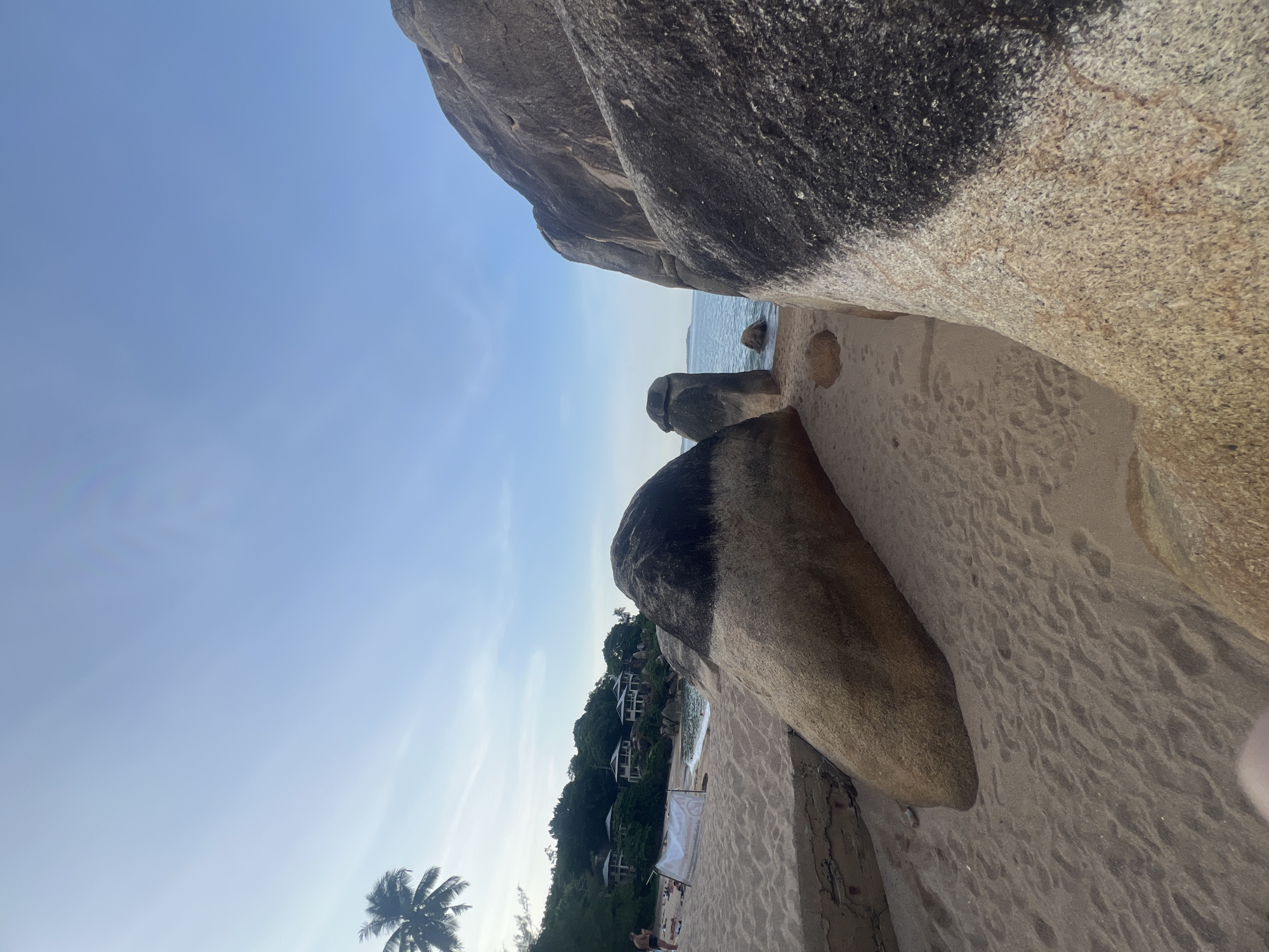 The image captures a beach scene in the Ko Samui District of Thailand. Dominating the foreground are large, weathered boulders, with their rough surfaces textured by sand and subtle color variations ranging from light tan to dark grey. The sand itself appears fine-grained and lightly rippled from the tide or wind. In the middle ground, more boulders are visible, some standing vertically like natural sculptures, separating the sandy expanse from the calm, blue ocean. The water appears clear and tranquil, with a slight shimmer under the bright sky.
Further in the background, lush green vegetation covers a hillside, interspersed with several modern, multi-story buildings, likely resorts or villas, featuring white walls and dark roofs. A tall palm tree with its fronds clearly visible stands to the left, its silhouette contrasting against the sky. On the beach, a few people are casually scattered, some appearing to be sunbathing or walking. A white banner with writing is propped up in the sand, its purpose unclear. The sky above is a pale, clear blue, with a hint of wispy clouds near the horizon. The overall atmosphere suggests a peaceful tropical day at a popular tourist destination.