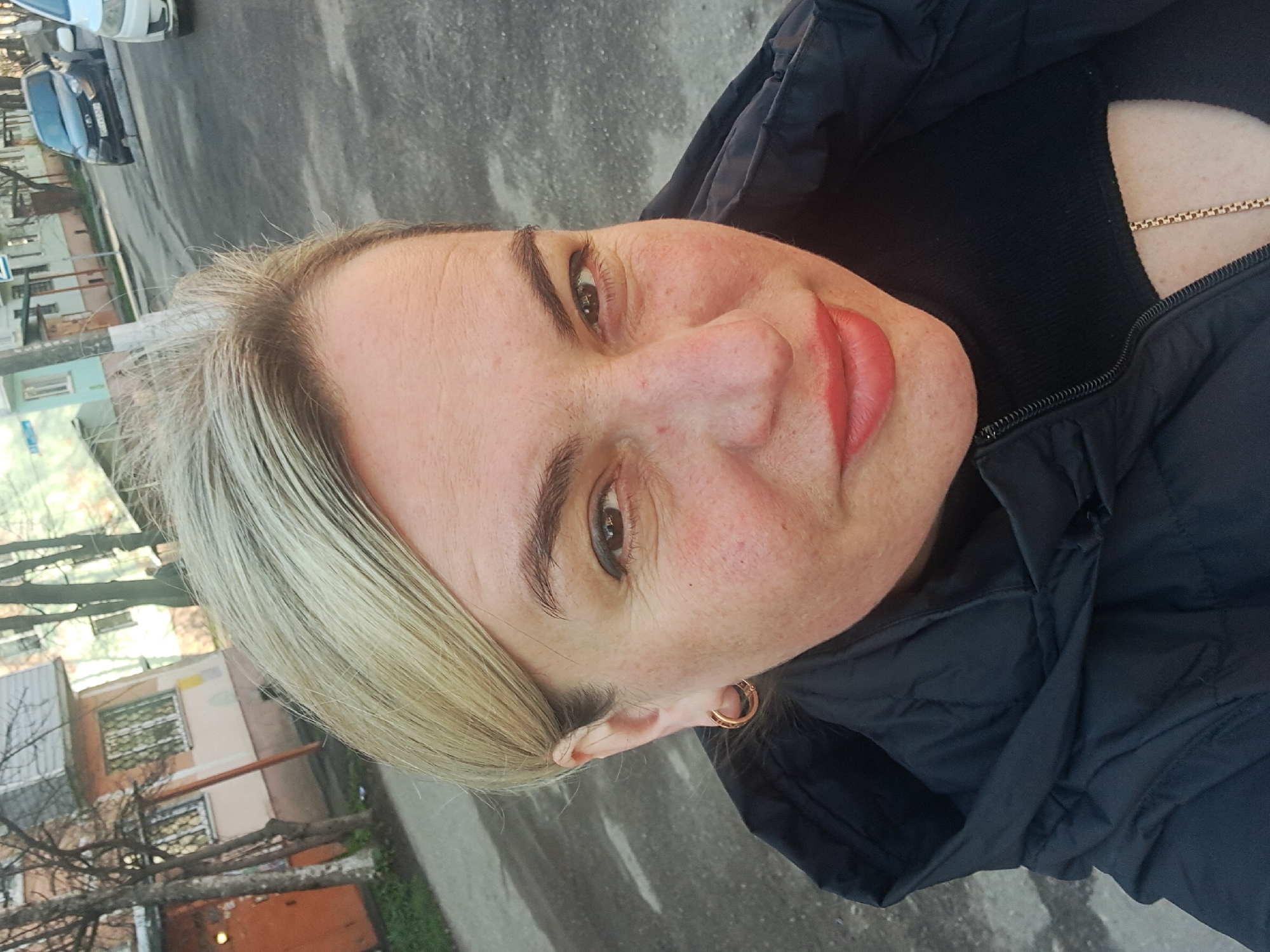 A woman with short, light blonde hair is taking a selfie. She is wearing a black jacket and a black top. A gold chain is visible around her neck. She has light skin, dark eyebrows, and is wearing lipstick. She is wearing gold hoop earrings.
The background shows a city street with buildings and cars. Based on the appearance of the buildings and the street, it is possible this image was taken in a city, such as Elektrostal, Russia. The weather appears to be overcast. Trees without leaves are visible.