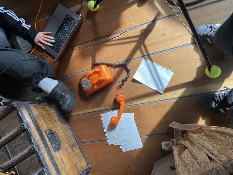 An overhead shot shows a wooden floor cluttered with items. There's a laptop, an orange rotary phone with the receiver off the hook, and scattered handwritten notes. A wooden chest is positioned near a person wearing black Crocs and a tracksuit. Another person is visible, wearing Adidas sneakers, and what appears to be a prop stand, stabilized by tennis balls. The scene is bathed in strong sunlight, creating distinct shadows.
