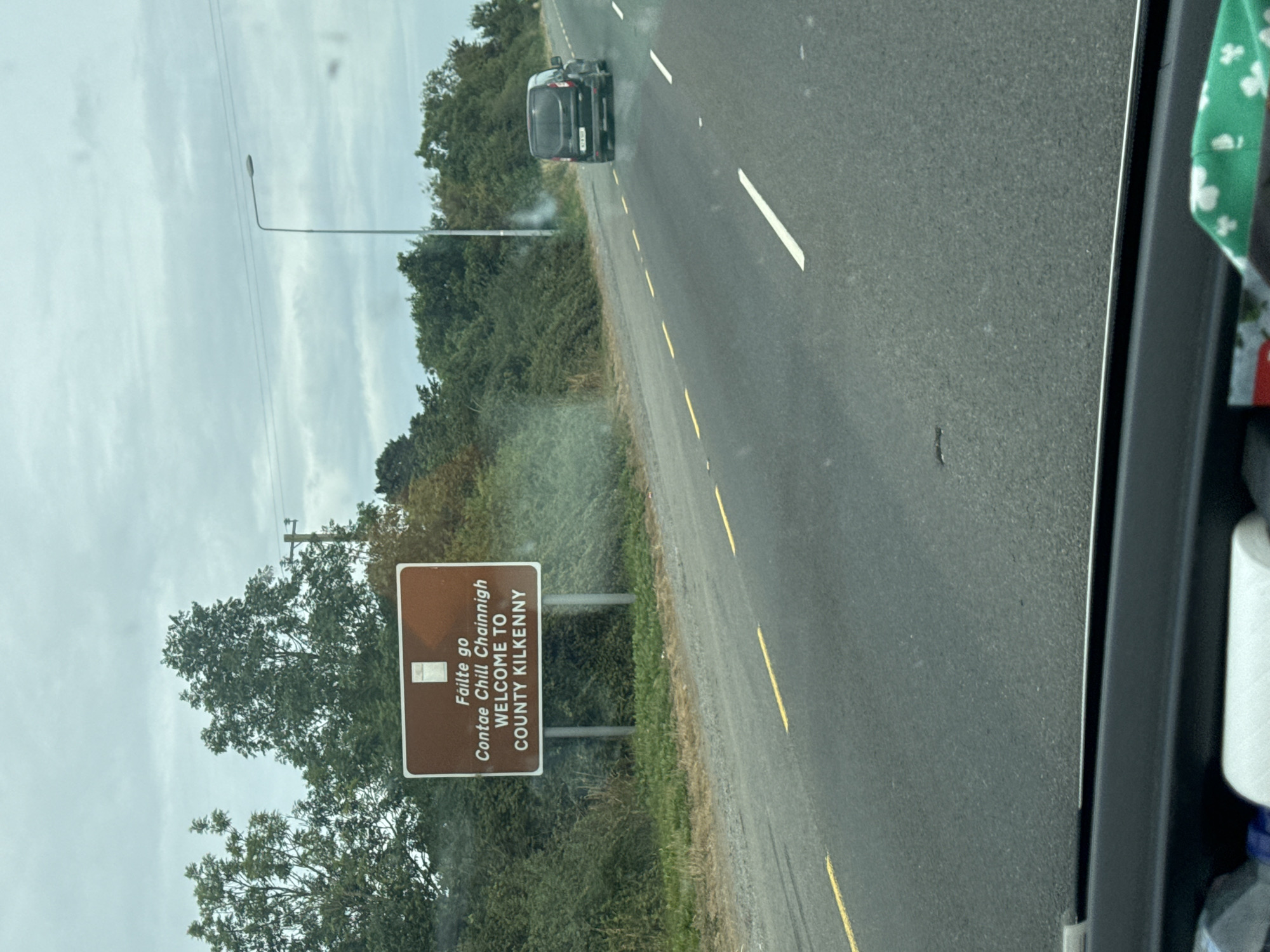 The image shows a brown sign on the side of a road that says "Fáilte go Contae Chill Chainnigh WELCOME TO COUNTY KILKENNY". A black van is driving on the road. There are trees and bushes on the side of the road, and the sky is cloudy. The location is County Kilkenny, Ireland.