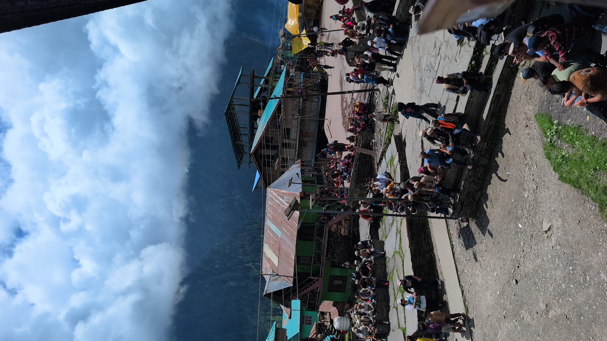 In Malana, India, a bustling gathering of approximately 100 people is captured on a paved, uneven courtyard. The scene is dominated by traditional Himachali architecture, with several multi-story wooden buildings featuring distinct corrugated metal roofs in various states of repair. One prominent green building displays a rusty, colorful roof and a set of stairs leading to an upper level. Adjacent to it, another building has a turquoise roof and balconies. Further in the background, a taller structure with multiple levels and a similar turquoise roof is visible, along with makeshift yellow awnings. The sky is dramatic, with large, soft white clouds obscuring a dark, forested mountain range in the distance.
The people are primarily seated on stone walls and steps that border the courtyard, while others stand or walk through the open spaces. Many are dressed in traditional Himachali attire, including distinctive caps. There's a sense of communal activity, with individuals engaged in conversations and observing their surroundings. Some are holding mobile phones, suggesting the presence of modern technology within this traditional setting. The ground is a mixture of stone paving, packed dirt, and patches of green grass. The overall impression is of a vibrant, communal gathering in a picturesque mountain village.