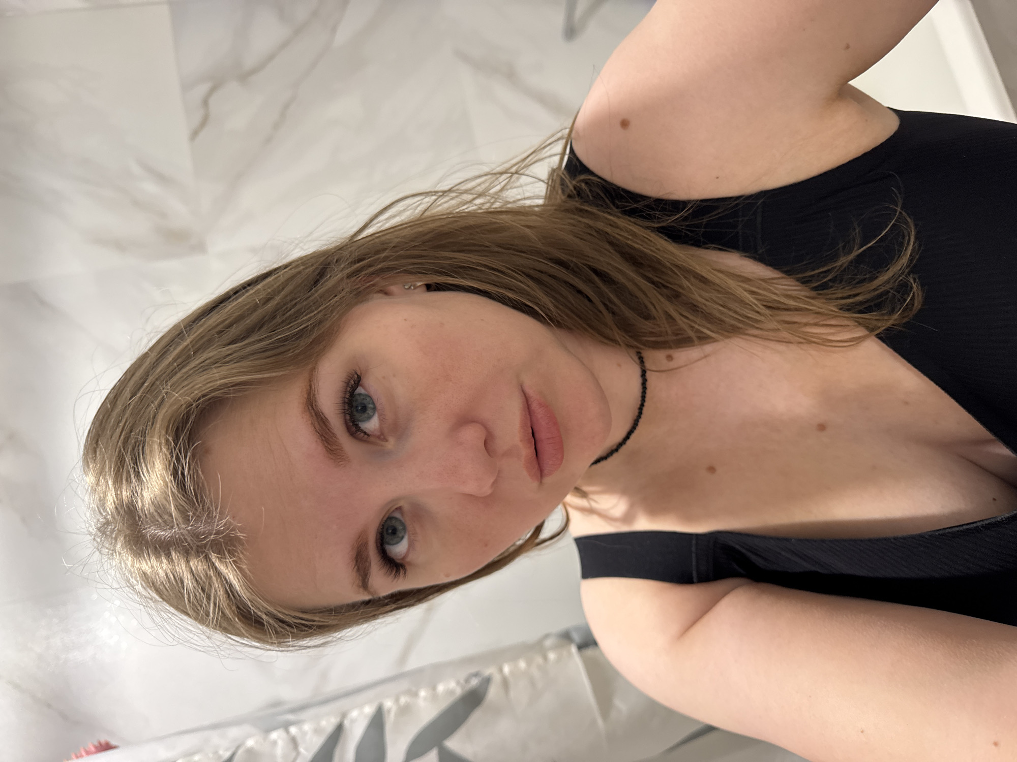 The image shows a woman with light brown hair and blue eyes in a black top, looking at the camera. She appears to be in a bathroom, evidenced by the marble-patterned wall and shower curtain in the background. The specific location is Ферма, Russia, but this information does not appear to be relevant to the scene depicted.