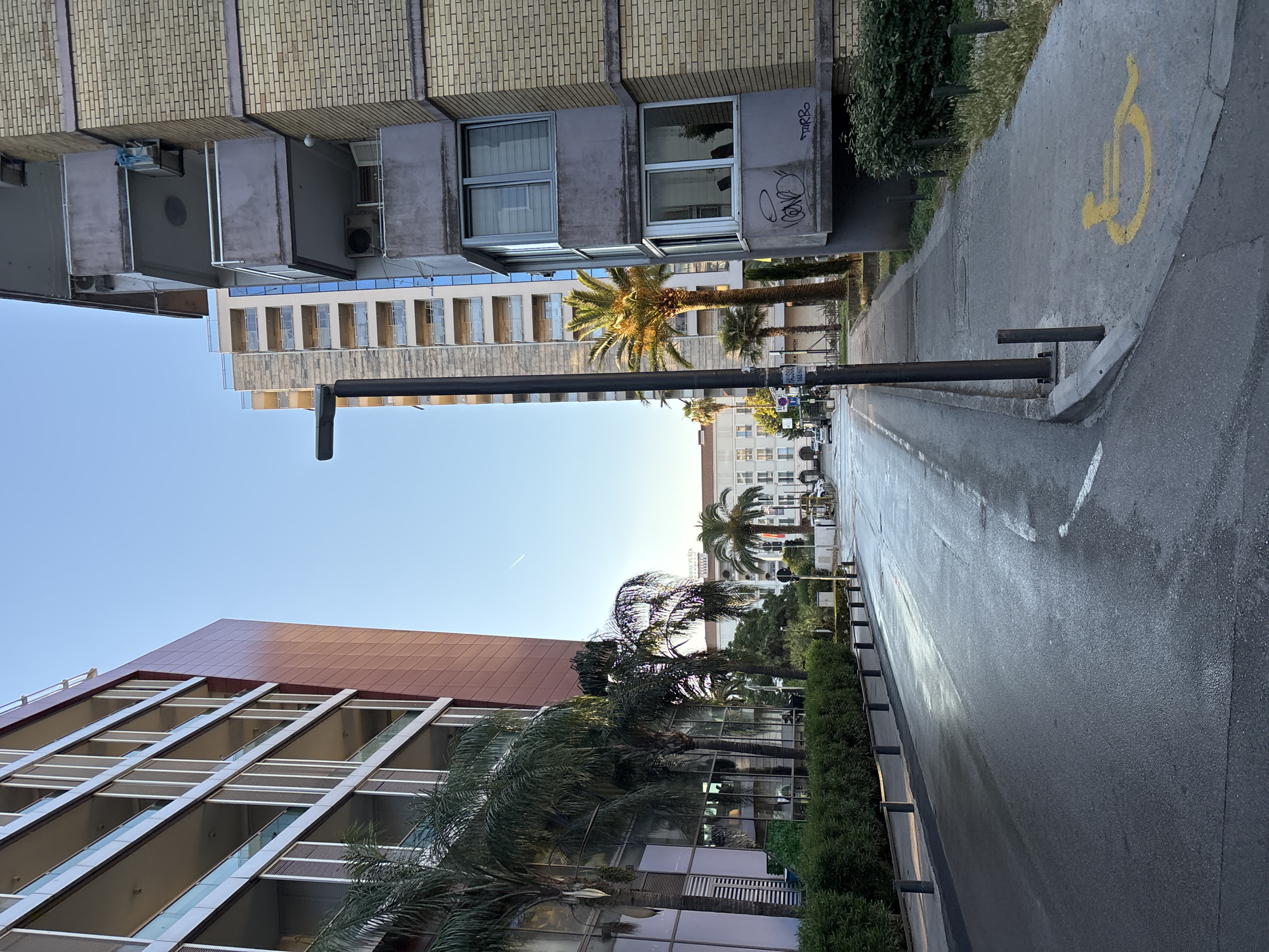 An eye-level, medium-shot captures a street scene in an urban environment, likely during the golden hour given the warm lighting on the buildings and road. On the left, a modern building with a reddish-brown facade and multiple balconies with glass railings dominates the view. Adjacent to it is a row of lush green bushes and palm trees. The road in front of these buildings is wet and reflects the light, suggesting recent rain or cleaning.
In the center of the image, a tall, dark street lamp stands on the sidewalk. Further down the street, more palm trees are visible, and behind them, several other buildings of varying architectural styles can be seen. The building on the right side of the frame is older, constructed with yellow bricks and featuring balconies with grey concrete trim. One balcony has graffiti sprayed on its underside. A yellow handicap symbol is painted on the concrete sidewalk in front of this building. The sky is clear and blue with a faint contrail from an airplane. The scene appears to be a quiet street with no people immediately visible.