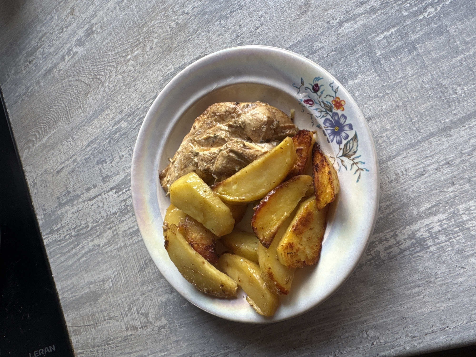 The image shows a plate of food, consisting of a piece of chicken and roasted potato wedges, placed on a textured gray surface. The plate is white with a floral pattern on the rim. A small portion of a black appliance is visible in the bottom left corner. The image was likely taken indoors under natural lighting. There is no information available to indicate the location is Chaikovsky, Russia.