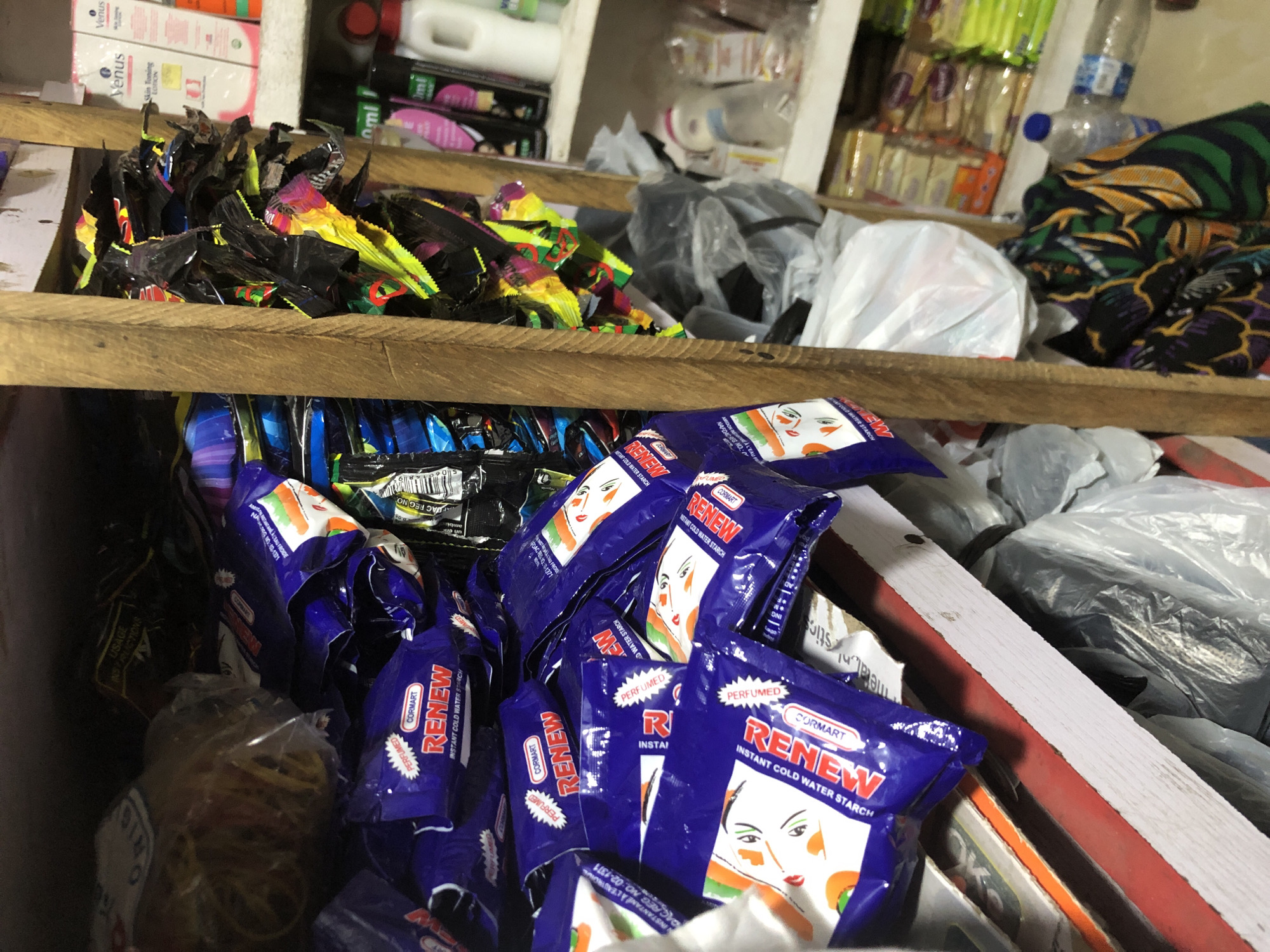 The image depicts a cluttered shelf in what appears to be a small shop or market stall, possibly located in Ado, Nigeria. The foreground is dominated by numerous purple packets of "RENEW INSTANT COLD WATER STARCH," some of which are piled high. These packets feature an illustration of a woman's face and brand markings. To the left of the starch packets, a plastic bag containing a bundle of yellow rubber bands is visible.
Behind the starch, a wooden divider separates the shelf into sections. To the right of the divider, a dark, chaotic pile of assorted packaging, primarily black with colorful accents, is crammed into a space. Further to the right, on a higher shelf, are various retail products. These include white and pink bottles of "Venus Skin Toning Lotion" and other cosmetic or personal care items. Below this, on another shelf, are stacked boxes of what appear to be food items or snacks, with prominent yellow and orange packaging.
On the lower shelves and floor area, more plastic bags, some crinkled and transparent, are piled up, along with a colorful, patterned fabric. A blue plastic bottle, possibly containing liquid, is also present on the bottom shelf. The overall environment suggests a busy, albeit unorganized, retail space. The lighting is dim, casting shadows and highlighting the textures of the various products.