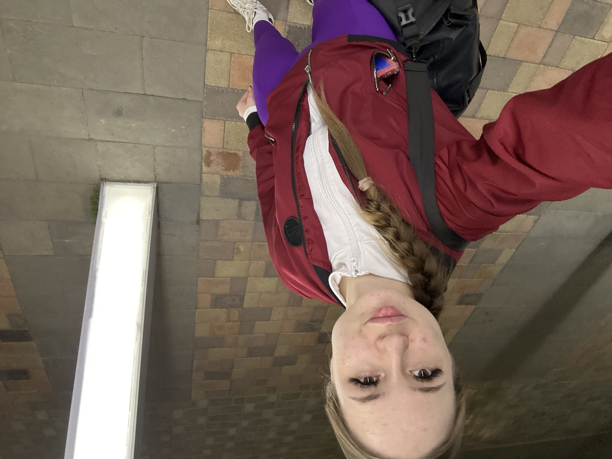 The image shows a young woman standing on a brick-paved surface. She wears purple leggings, white sneakers, a white top under an open red jacket, and a black backpack. Her blonde hair is braided. A long, rectangular light fixture is affixed to a wall on the left. The location appears to be an indoor space, possibly a walkway or station. The city is Unknown, Unknown.