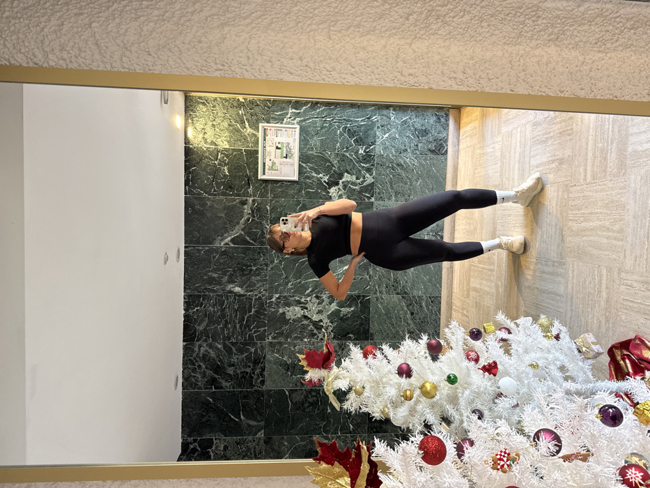 The image displays a full-body mirror selfie taken by a female individual in an indoor common area within Cannes, France. The woman is centered in the reflection, standing with her left hand on her hip and her right hand holding a light-colored smartphone towards the mirror. She is wearing a black short-sleeved crop top, black athletic leggings, white socks with a visible Nike swoosh, and light-colored athletic shoes. Her hair is light brown with bangs, and she wears eyeglasses or sunglasses and earrings.
The background reflected in the mirror consists of a wall covered in rectangular panels of dark green, possibly marble, tile. The ceiling is white with two recessed light fixtures. The floor is tiled with light-colored square tiles. To the left of the woman's reflection, two artificial white Christmas trees are prominently featured, adorned with a variety of red, gold, purple, and green spherical ornaments, along with other decorative elements such as gingerbread men and snowflakes. A large red and gold decorative leaf is visible at the top left above the trees. A red fabric covers the base of the Christmas trees. To the right of the woman's reflection, mounted on the green tiled wall, is a white framed emergency evacuation map with a red border, labeled "PLAN D'ÉVACUATION" at the top. Outside the mirror's frame on the far right, a textured, off-white wall surface is visible.