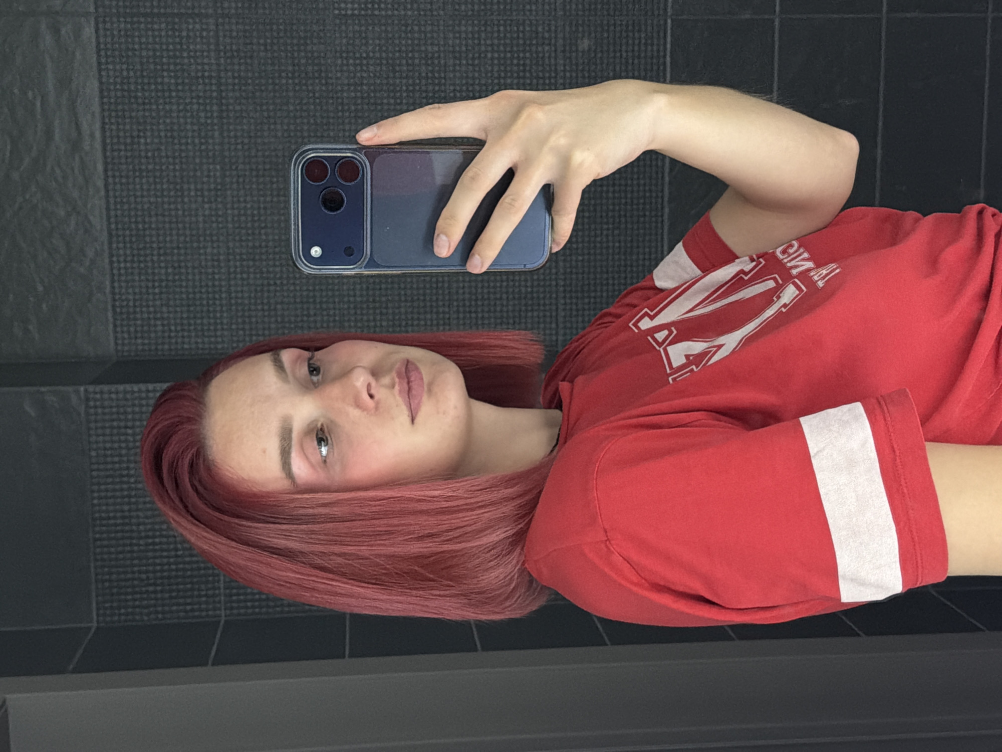 A young woman with red hair is taking a selfie in a bathroom. She is wearing a red t-shirt with white stripes on the sleeves and a white logo on the chest. The bathroom has dark tiled walls. The woman is holding a blue phone in her right hand. The image is taken from the chest up.