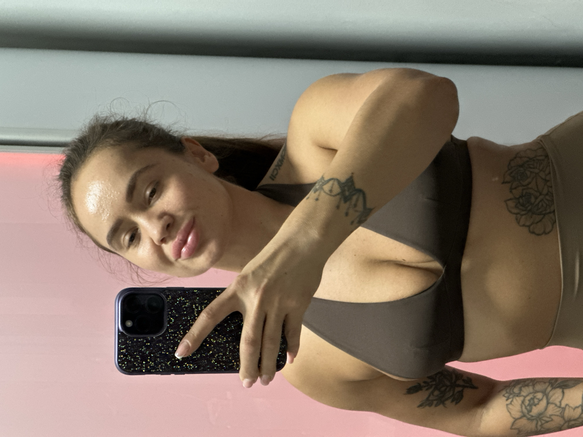 A woman with dark hair pulled back is taking a selfie in a room with a pink wall on the left and a gray wall on the right. She is wearing a brown bikini top and tan bottoms. She has several tattoos on her arms and torso, including floral designs and what appear to be Roman numerals on her shoulder. She is holding a black phone with a sparkly case in her right hand, angled to capture her reflection. Her lips are slightly parted, and she is looking at the phone's screen. The lighting in the room appears to be bright, casting some glare on her forehead.