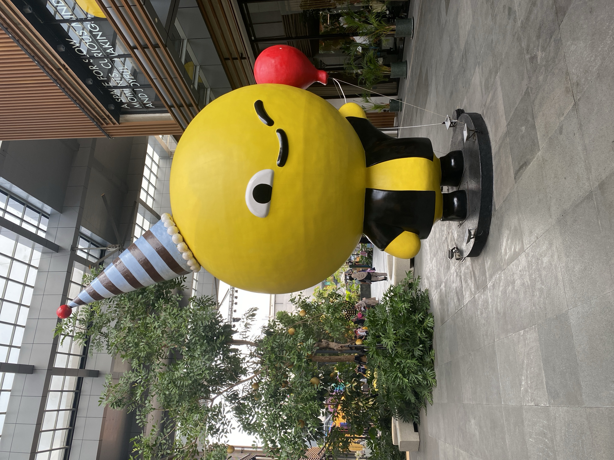 The image depicts a large, yellow, bee-like cartoon character statue wearing a black and yellow striped outfit and a blue and brown striped party hat. The statue is holding a red balloon in its right hand and has a winking expression. The statue is situated in an open, paved area, possibly a plaza or walkway, with modern architecture in the background. To the left of the statue, there is a tree with yellow spherical fruits and lush green foliage. Several people are visible in the background, walking around and interacting in what appears to be an outdoor commercial or recreational space. The overall environment suggests a vibrant and lively atmosphere. This scene is located at PIK Avenue Mall in North Jakarta, Indonesia, a prominent shopping and lifestyle destination.