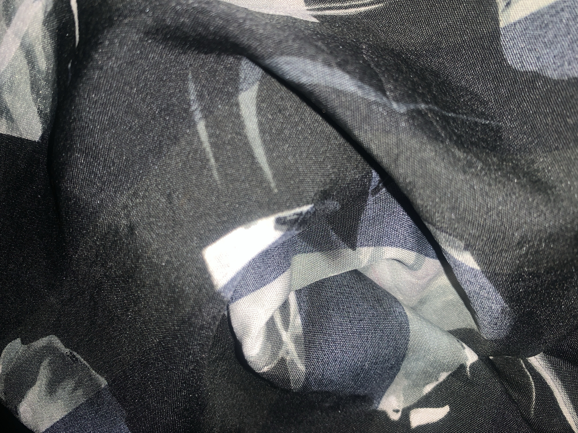The image shows a close-up view of a wrinkled, dark-colored fabric with a pattern of abstract shapes in shades of white, gray, and blue. The fabric appears to be synthetic and has a slightly reflective sheen. There are no visible people, animals, or specific landmarks in the image. The lighting suggests an indoor setting or a shaded outdoor area. The image does not contain any information that can be used to determine its location in Jalingo, Nigeria.