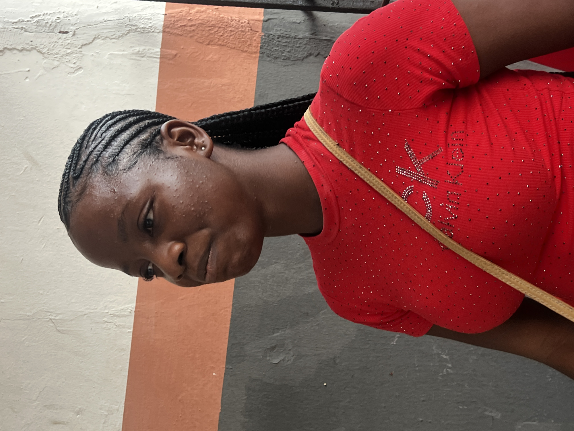 A medium shot captures a young Black woman with braided hair from the chest up, standing in front of a textured wall. The wall is divided horizontally into three distinct color bands: a pale off-white at the top, a burnt orange in the middle, and a dark gray at the bottom. The woman is looking to her left, her expression neutral, with a slight pursing of her lips. She has small earrings in her ears and several small blemishes on her cheeks and forehead. Her hair is styled in thin cornrows that extend into long braids down her back. She is wearing a bright red, short-sleeved t-shirt adorned with numerous small, shimmering embellishments, possibly rhinestones or sequins, that form a subtle pattern. A thin, tan strap, possibly from a bag, crosses her chest diagonally from her left shoulder to her right side. The image appears to be taken outdoors, with the lighting suggesting daytime. The background wall has some visible wear and tear, with patches of discoloration and cracking, suggesting an urban environment. The location is identified as Nkpor, Nigeria.