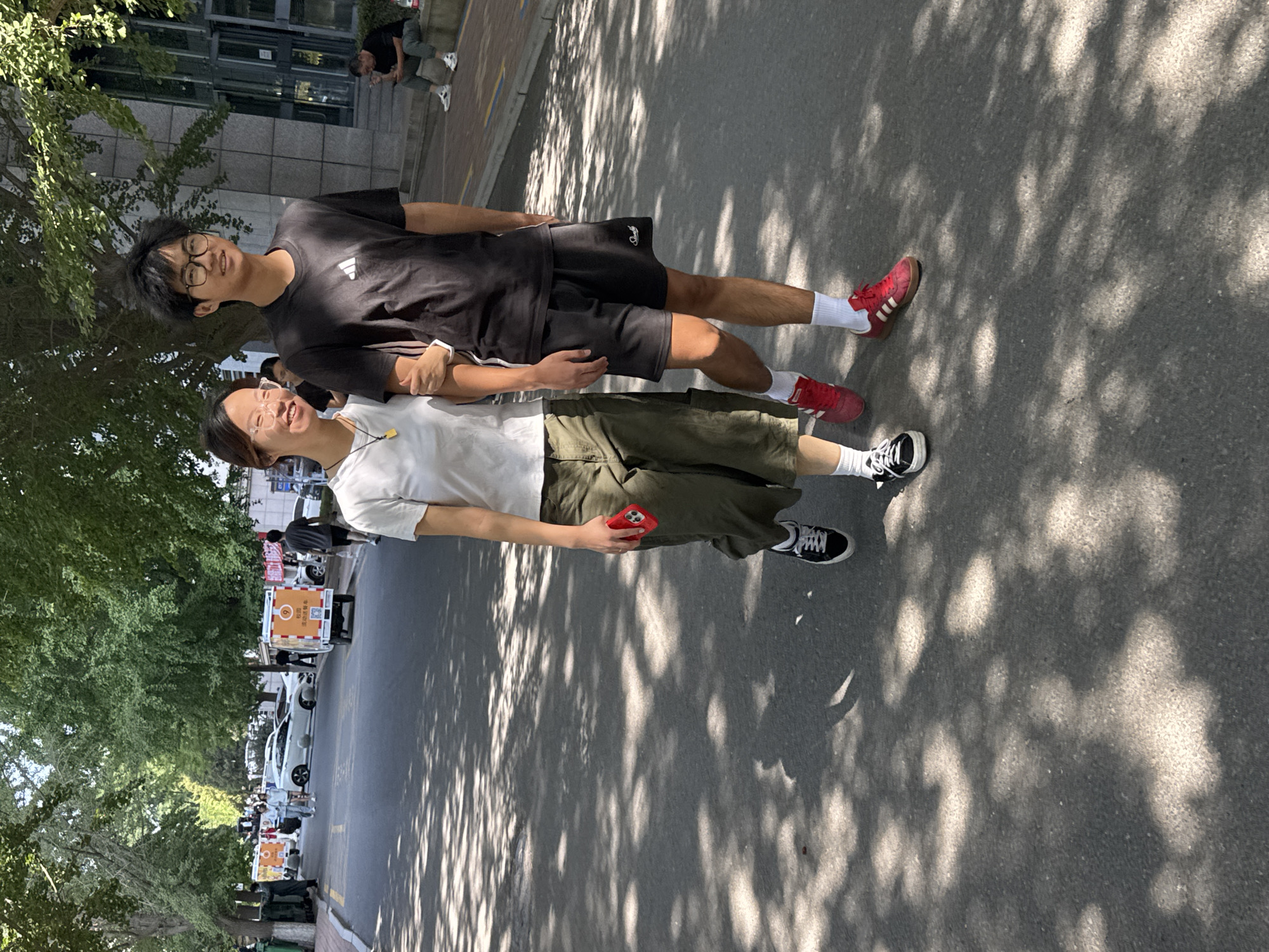 In the city of Zhongtun, China, a young Asian couple walks arm-in-arm on an asphalt road partially shaded by overhead trees. The female, on the left, wears a white t-shirt, olive green cargo pants, and black and white sneakers, holding a red-cased smartphone in her right hand. The male, on the right, sports a black t-shirt with a white Adidas logo, black shorts, white socks, and red sneakers. In the background, a white vehicle with orange branding and a covered cargo box is parked on the side of the road, and several other vehicles and pedestrians are visible further down the street. A building with a modern facade is situated to the right of the path, where a man is seated on a low wall. The sunlight casts dappled shadows from the trees onto the road.