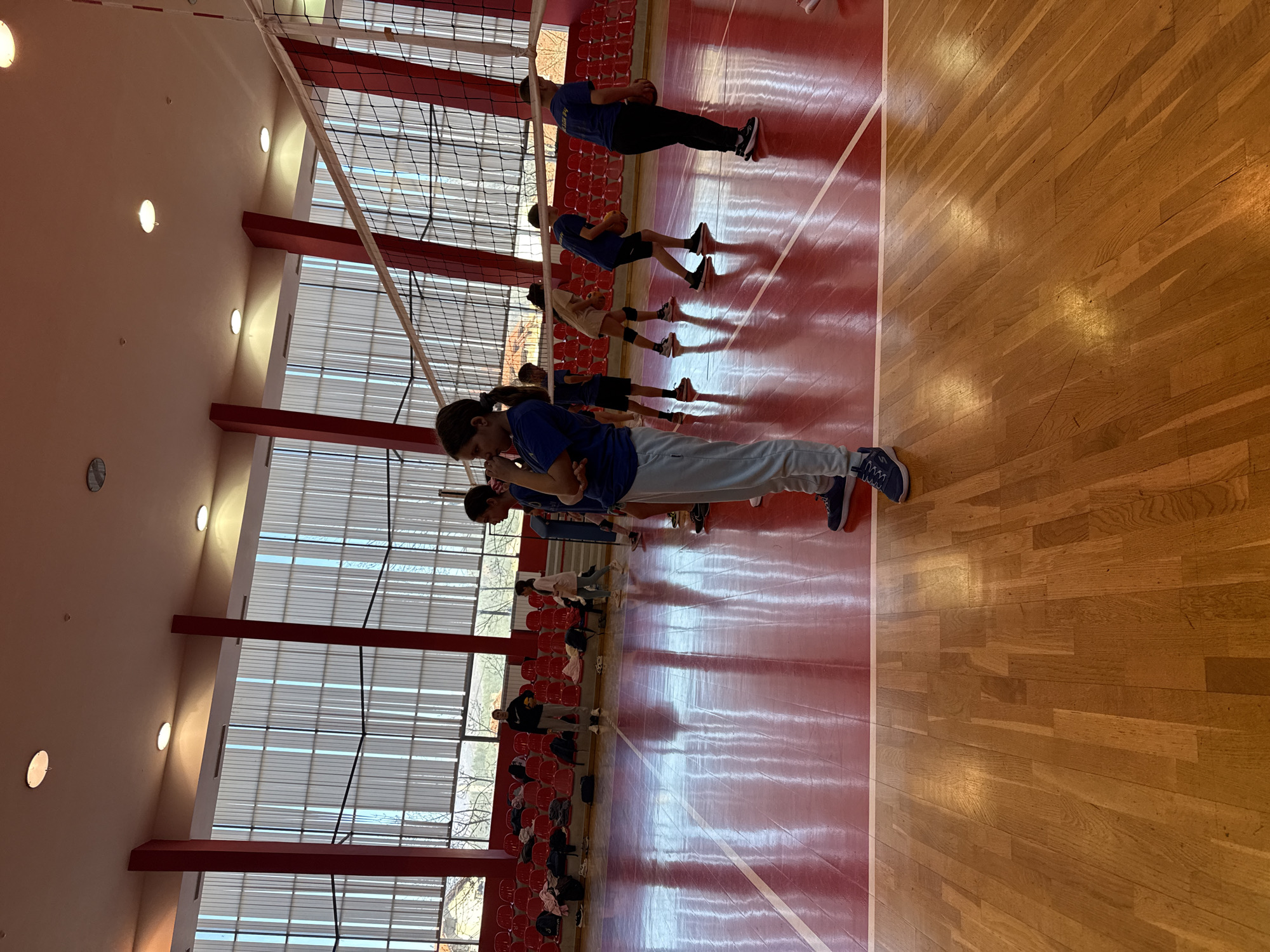 A gymnasium in Etropole, Bulgaria, features a wood floor transitioning to red near the wall and red seating. A group of young people in blue shirts stand in a line, some holding balls, suggesting a practice or drill. Large windows illuminate the space, showing trees outside.