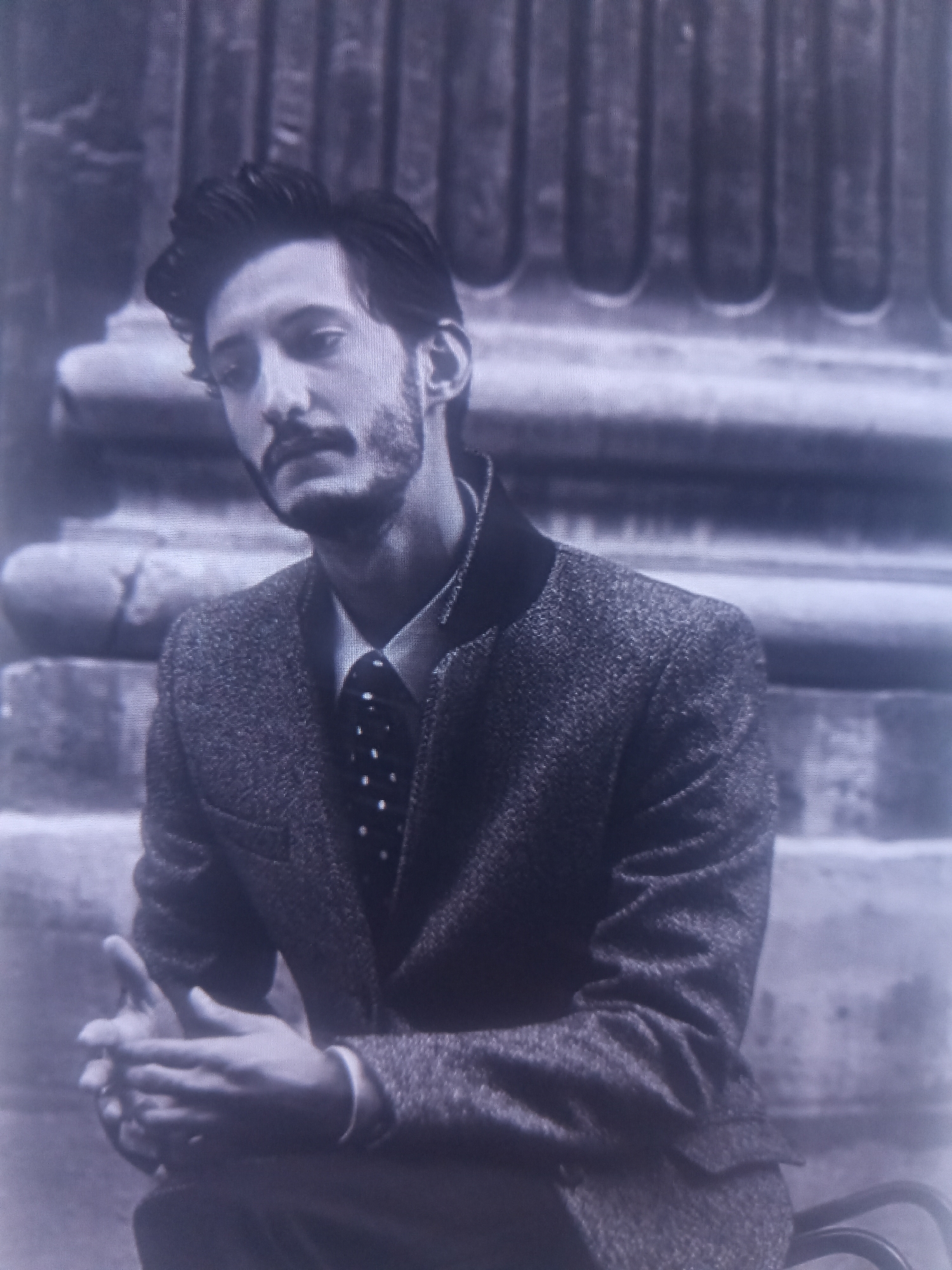 A black and white, medium shot, eye-level, outdoor portrait shows a man with dark hair, a mustache, and a beard, leaning forward with his hands clasped in front of him. He is wearing a dark tweed jacket with a black collar, a white collared shirt, and a dark tie with white polka dots. The man is seated in front of a textured, stone architectural element, possibly a column or facade, with a blurred background. The lighting is soft and diffused, creating a slightly grainy and atmospheric effect.