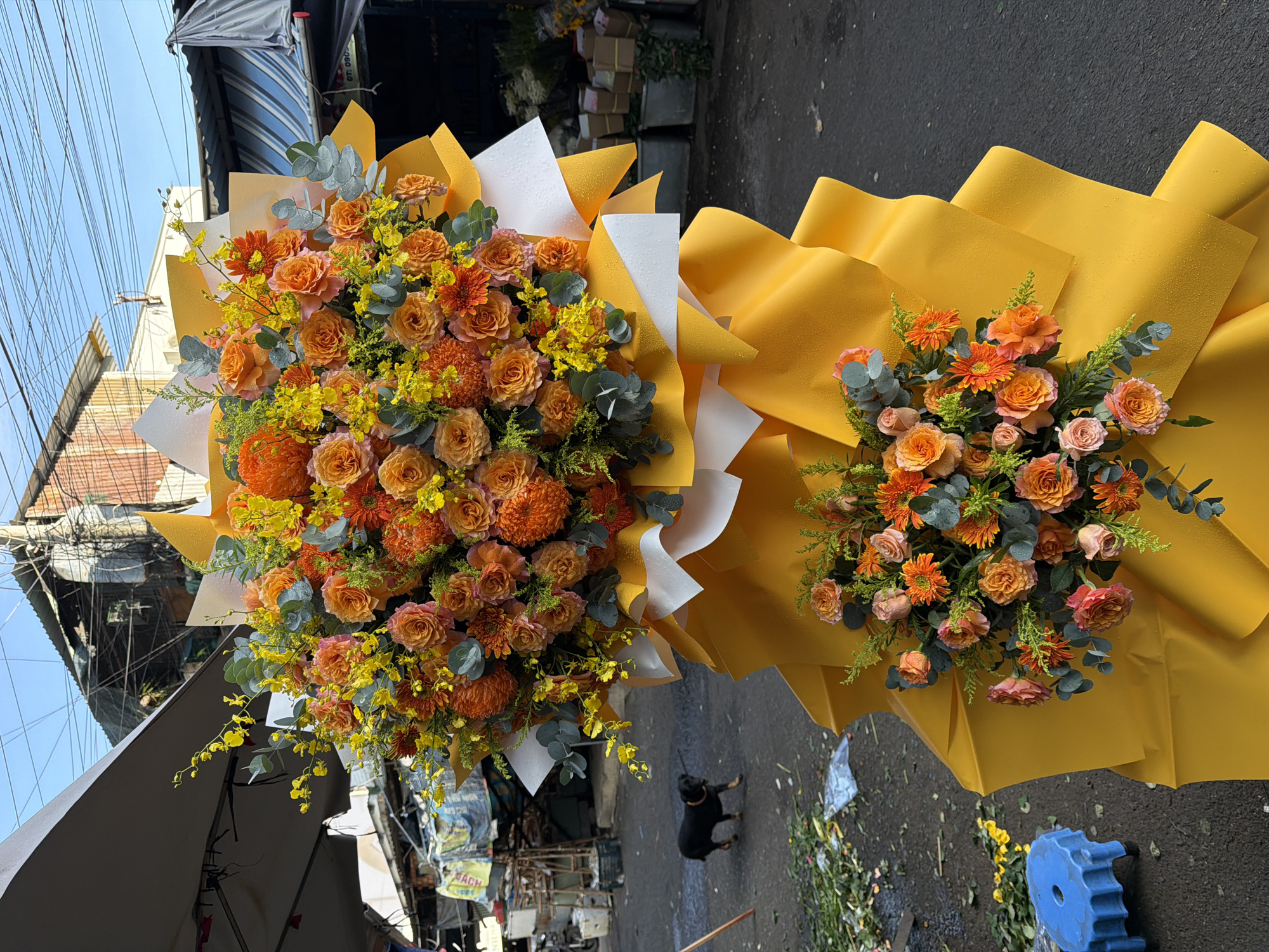 This image depicts a vibrant floral arrangement, likely a standing spray, adorned with numerous orange and yellow roses, chrysanthemums, and smaller yellow flowers, accented with eucalyptus leaves. The arrangement is presented in a bold, layered yellow wrapping, with white paper visible beneath the top section. The scene appears to be outdoors, possibly at a flower market in Nha Trang, Vietnam, given the context of the street, market stalls with striped awnings, and buildings in the background. A small black dog with a red collar is walking on the paved ground in the mid-left of the frame. Discarded flowers and debris are scattered on the ground around the arrangement and to the left. A blue plastic stool is visible in the lower-left corner. The overall impression is one of a lively, possibly bustling, outdoor marketplace.