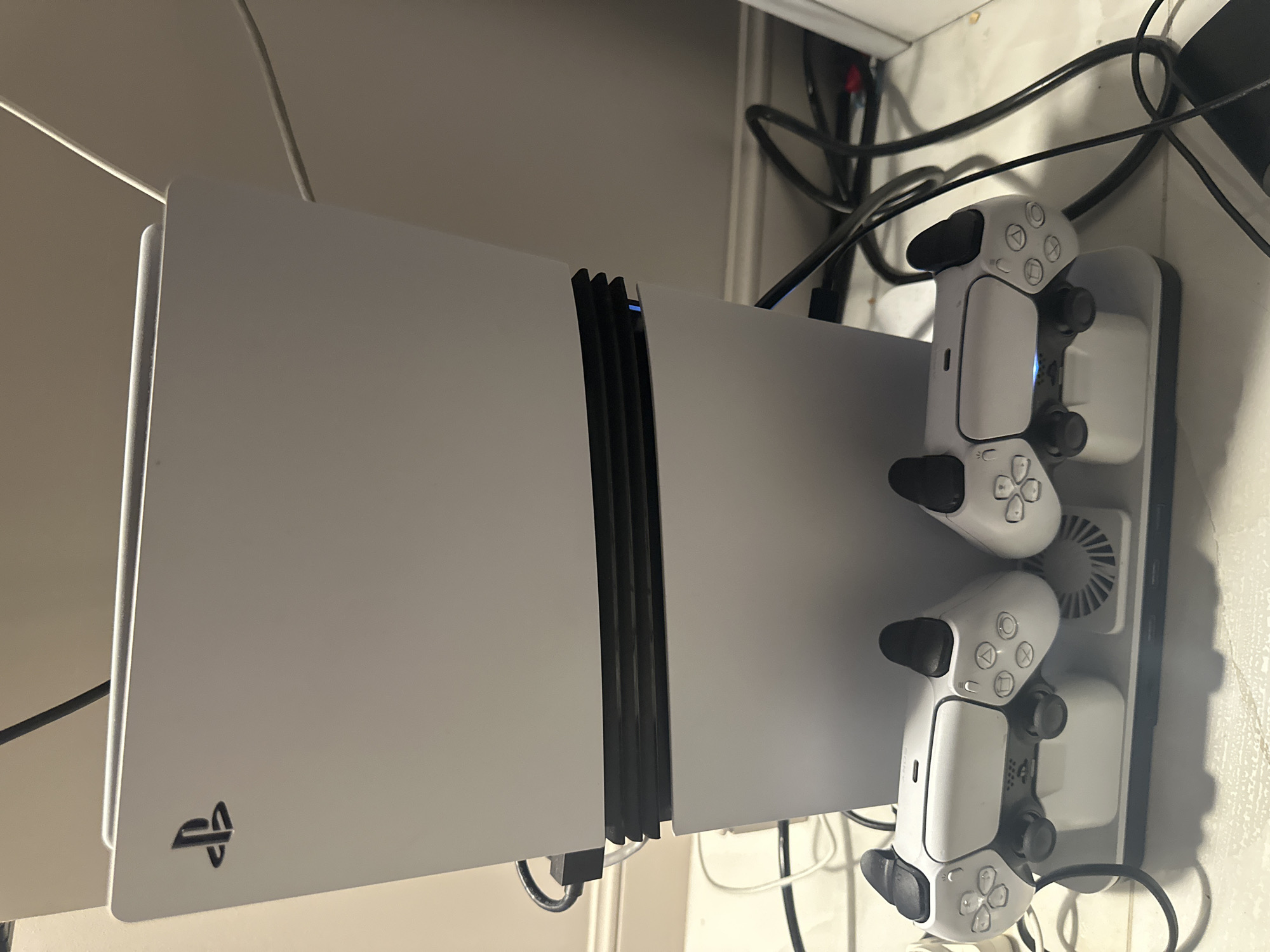The image displays a modern gaming setup featuring a white PlayStation 5 console and two white PlayStation DualSense controllers, situated in an indoor environment in Moscow, Russia. The PlayStation 5 console stands vertically on a surface, likely a desk or shelf. A subtle blue light emanates from the central black vent of the console, indicating that it is powered on. The stylized PlayStation logo is visible on the lower left portion of the console's faceplate.
Adjacent to the console, two PlayStation DualSense controllers are docked in a black charging station. The controllers exhibit their characteristic white design with black joysticks and trigger buttons. Visible text on the controllers includes the iconic PlayStation button symbols: triangle, circle, X, and square, as well as the PlayStation logo on their touchpads. The charging station also features a circular fan grill, suggesting it may provide cooling for the console or controllers. The background is filled with a network of black and white cables, characteristic of electronic equipment, against a light-colored wall or furniture. The scene captures a neat but cable-dense gaming station.