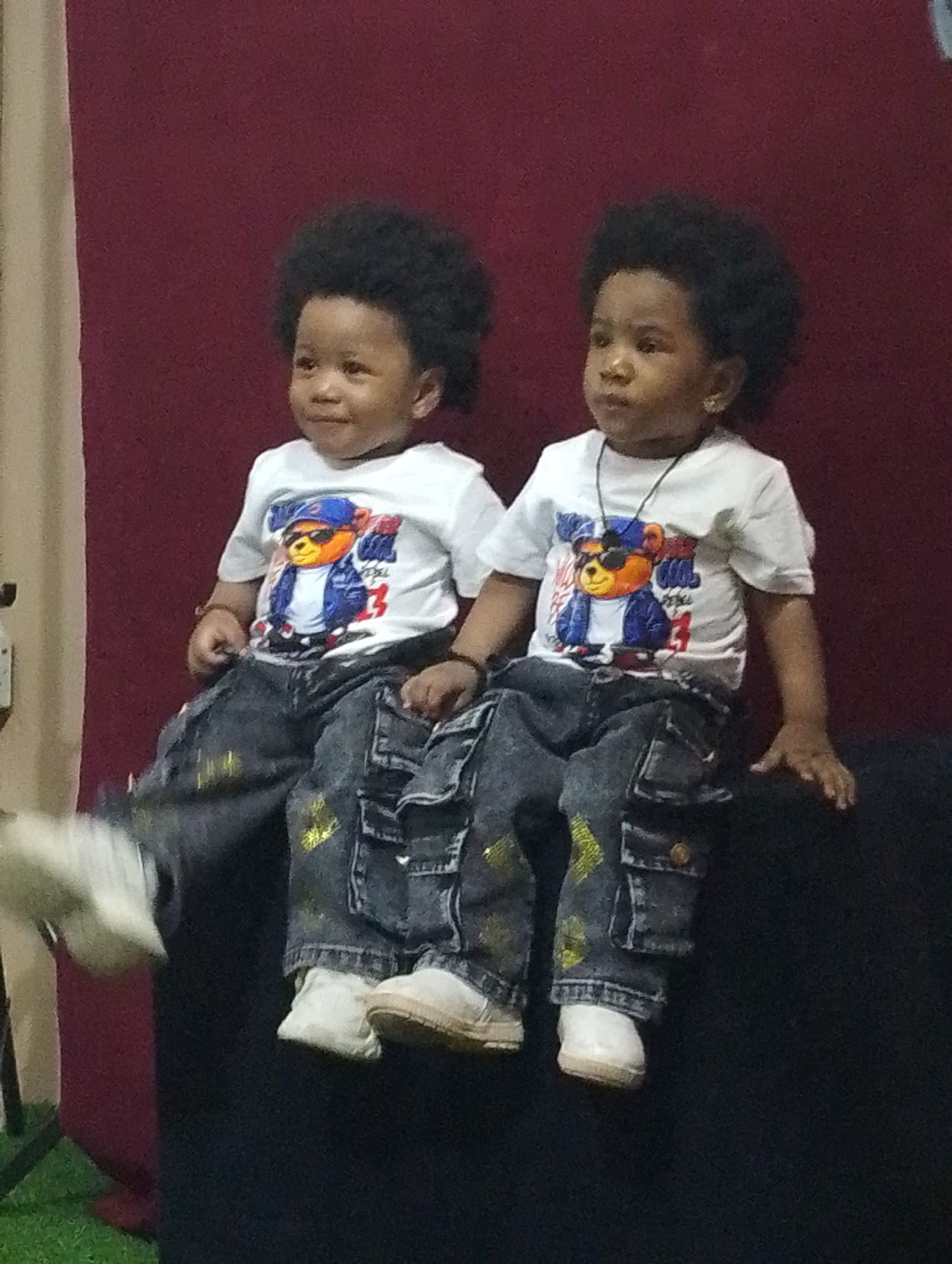 Two young children, likely twins or siblings, are seated side-by-side against a plain reddish-brown or maroon backdrop in what appears to be an indoor setting in Babban Giji, Nigeria. Both children have dark, curly hair and dark skin. They are dressed in matching outfits: white short-sleeved t-shirts featuring a graphic of a cartoon bear wearing sunglasses, a blue jacket, and a blue cap, along with dark blue jeans adorned with yellow or gold paint-splatter designs. Each child also wears light-colored, possibly white, shoes.
The child on the left looks slightly to their right with a soft smile. The child on the right looks off to the right with a more neutral, inquisitive expression; this child also wears a thin necklace with a small pendant and a small stud earring in their right ear. The child on the left appears to have a bracelet on their left wrist. A faint red number "3" is visible on the right side of the bear graphic on the t-shirt worn by the child on the right. On the left side of the image, a vertical light-colored element, possibly a curtain or wall, is visible, along with a patch of green at the bottom. The children appear to be posing for a photograph.