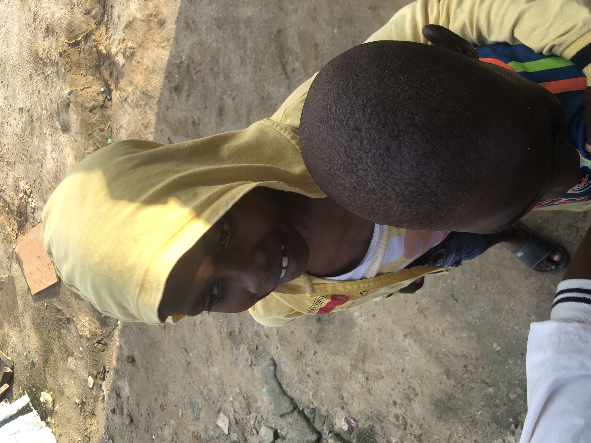 In Ado, Nigeria, a high-angle shot captures two young children, one wearing a yellow hooded sweatshirt and a white t-shirt with a colorful stripe pattern visible underneath. The child in the yellow hoodie has their hood up, revealing their face which is turned slightly to the left, and they are smiling, showing teeth. Their left arm is extended, and their right arm is bent. The second child, mostly seen from the back of their head, has dark skin and is wearing a striped t-shirt. The ground beneath them is a rough, dusty surface with scattered debris and small rocks. The scene appears to be outdoors in what could be a semi-urban or rural setting, with muted lighting and shadows suggesting daylight. A white leg with black and white striped socks is visible at the bottom left of the frame.