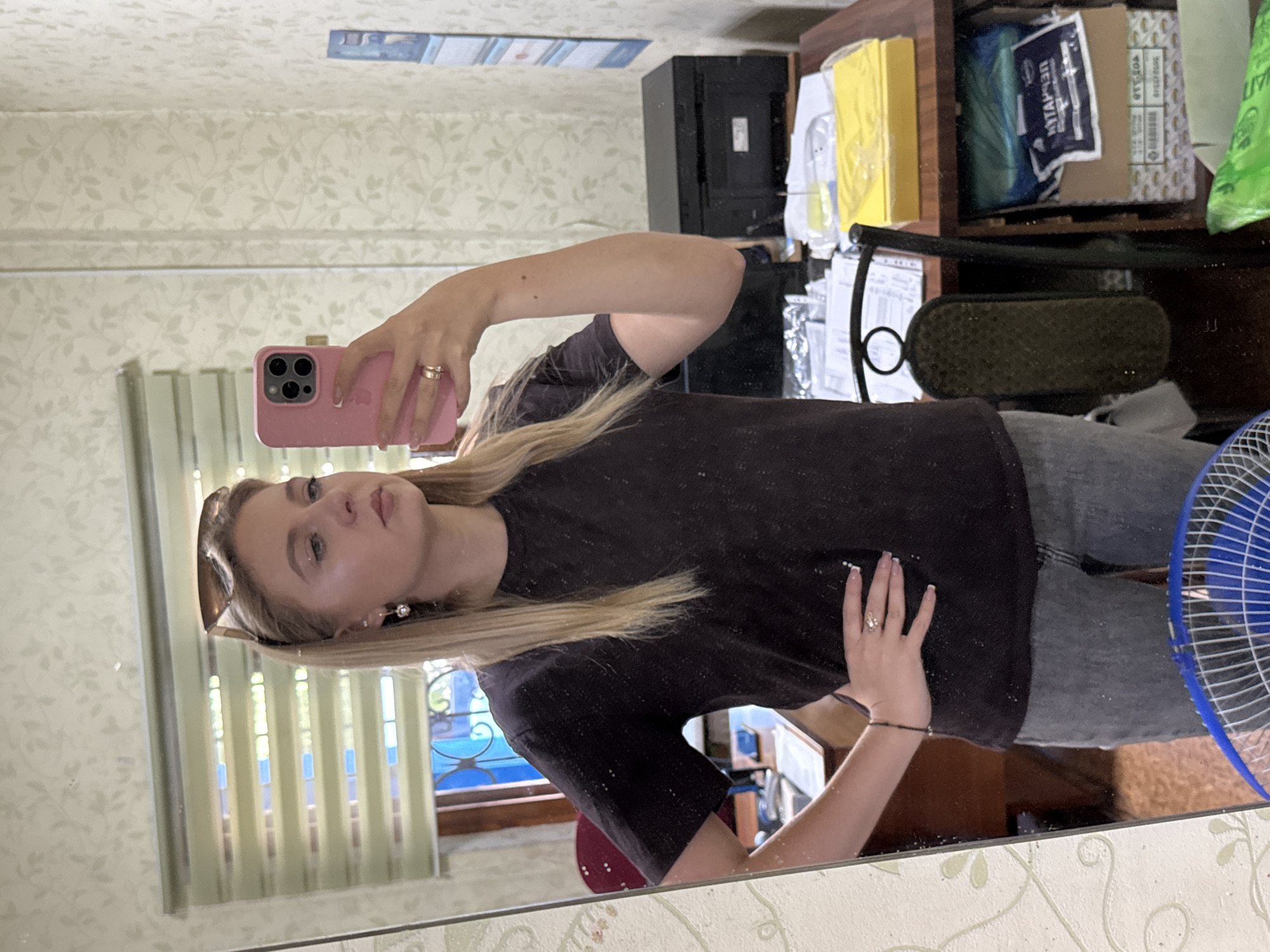 A young woman with blonde hair and sunglasses on her head is taking a mirror selfie in what appears to be a home office. She is wearing a dark t-shirt and grey jeans. She has a pink phone in her right hand. The background shows a desk with a printer, papers, and various office supplies, as well as blinds on a window. The room is decorated with floral wallpaper.