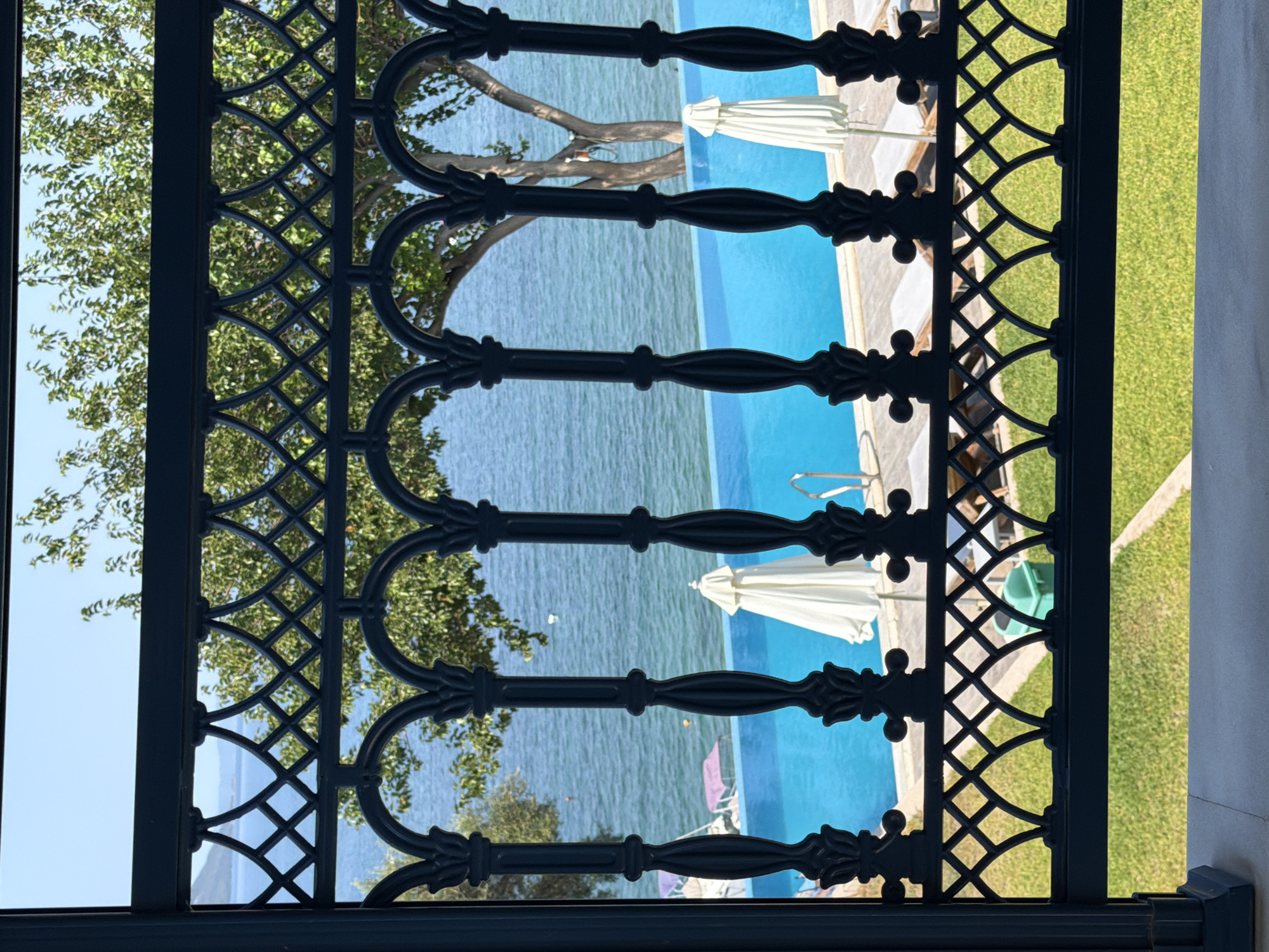 The image shows a view through a decorative black metal railing. Beyond the railing, a bright blue swimming pool is visible, with lounge chairs and white umbrellas positioned around it. The sea stretches out in the background, with a few boats in the distance. Trees with green foliage are also present, and the sky is clear and blue. The foreground shows a patch of green grass. The scene appears to be a resort or hotel in a sunny, coastal location. The presence of the sea, pool, and lounge chairs strongly suggests a vacation or resort setting. The ornamental railing indicates a decorative architectural style. The overall impression is one of a relaxed, leisurely environment.