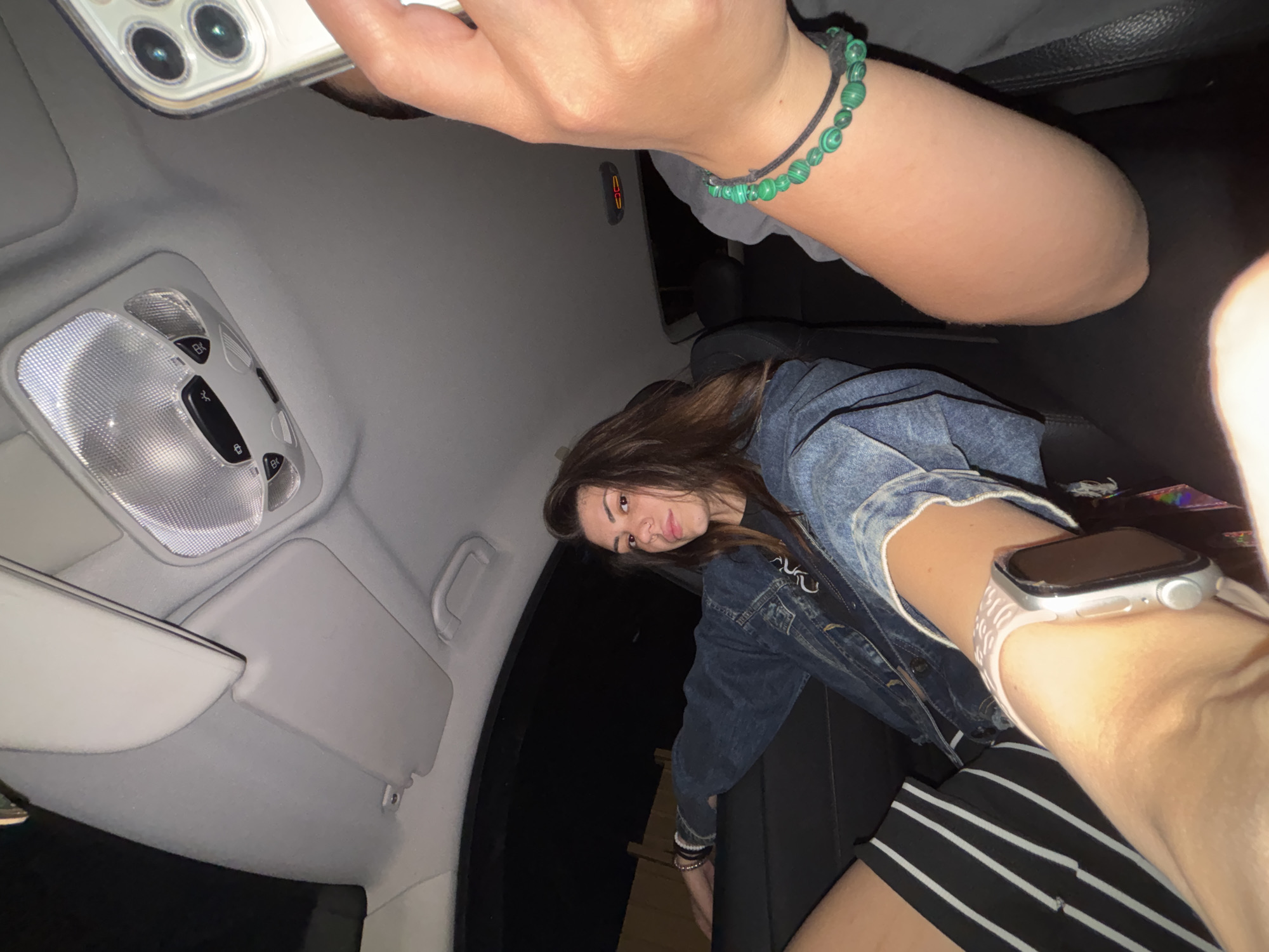A wide-angle interior photograph captures two individuals within a modern vehicle, located in Sofia, Bulgaria, as indicated by the prompt. The scene is illuminated by interior lights and appears to be set at night or in a dimly lit environment.
In the left foreground, the left forearm of a young adult female is visible, adorned with a silver-bodied Apple Watch with a white band. This individual is the primary subject, seated in the rear left passenger seat. She possesses long brown hair, a nose piercing, and is dressed in a blue denim jacket layered over a dark t-shirt with white graphic elements, and black and white striped shorts. She looks directly at the camera with a composed expression.
To her right, partially visible, is the right arm and hand of a second individual. This person is holding a silver or white iPhone (featuring a prominent triple-lens camera array) in a clear protective case, presumably taking the photograph. Their right wrist is accessorized with a green beaded bracelet and a black cord bracelet.
The car's interior features a light gray headliner and a detailed overhead console. The console contains a textured clear dome light, control buttons with various symbolic labels (suggesting functions like interior lighting and possibly ventilation/sunroof controls), and a microphone grill. A matching light gray sun visor is partially visible in the folded-up position, and a grab handle is present above the main subject. The windows reveal a dark exterior, consistent with nighttime conditions. The design and layout of the overhead console strongly resemble those found in Mercedes-Benz vehicles of a certain generation.