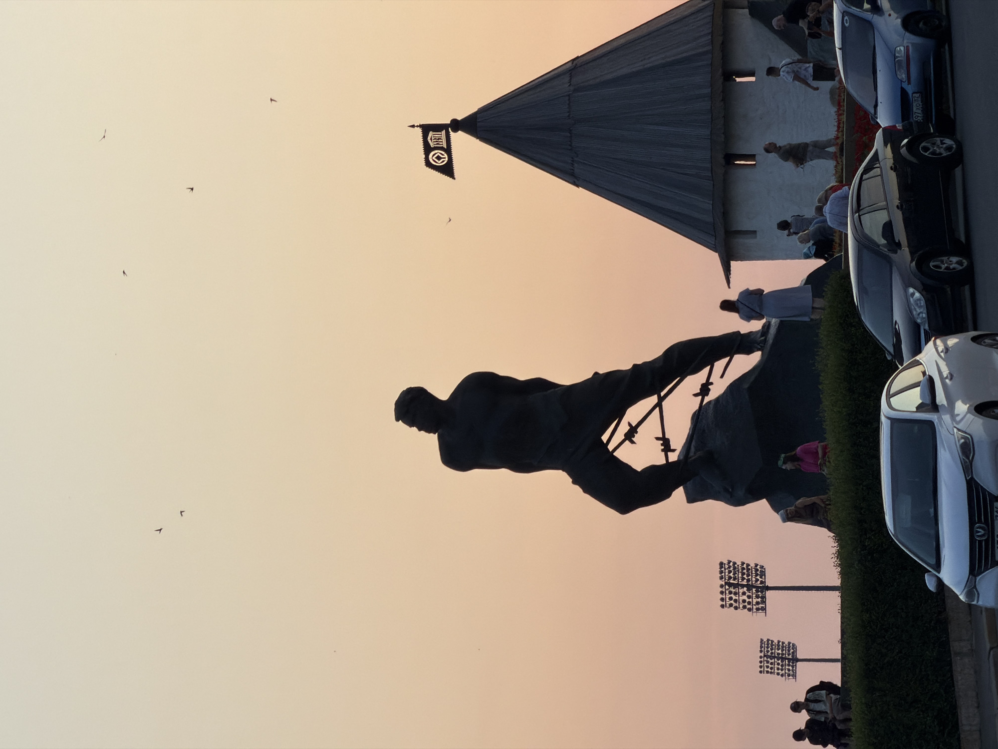 The image depicts a silhouetted statue of a male figure, possibly a worker or athlete, dynamically posed against a sunset sky with hues of orange and pink. The statue stands on a rocky base, with some barbed wire-like elements visible. To the right of the statue is a conical-shaped building, resembling a tower or a section of a fortress wall, with a dark flag featuring an emblem at its peak. Several people are scattered around the base of the tower and in the foreground, some sitting on a low hedge, others standing and conversing. In the background, two tall light poles with numerous lamps are visible. In the foreground, three cars are parked on a paved surface, with one white car in the immediate foreground. The overall scene suggests an outdoor public space, likely a park or a historical monument site. This scene is identified as the monument to Musa Dzhalil in Kazan, Russia.