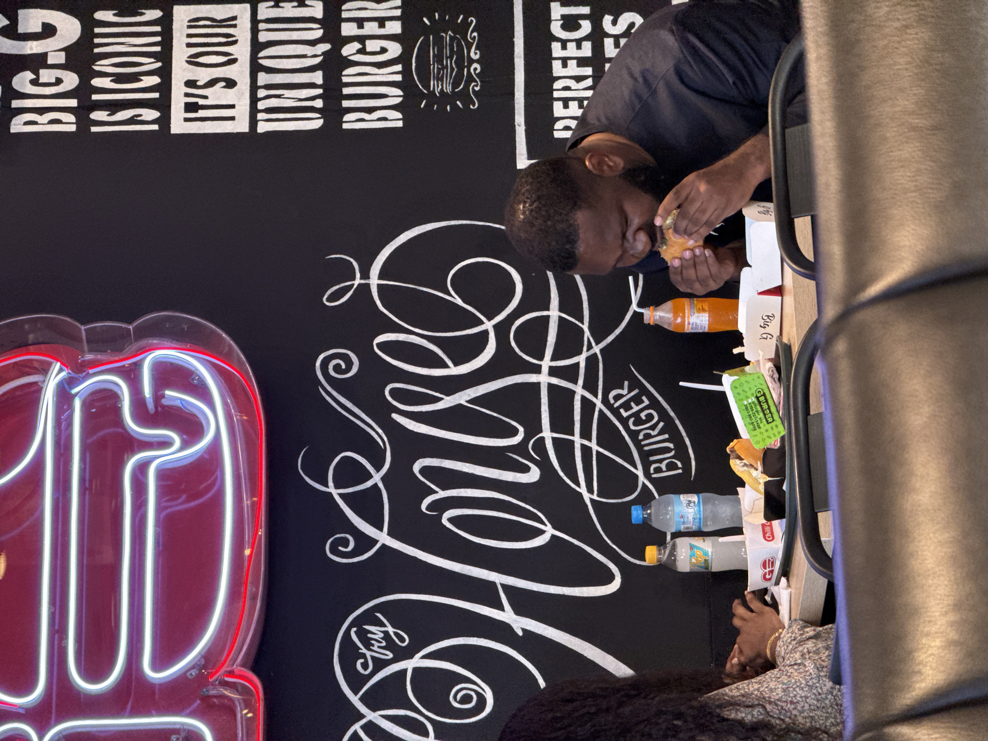 The scene depicts the interior of a restaurant, likely in Abuja, Nigeria, based on the branding "Big G Burger" and associated phone numbers. A man is seated at a table, eating a burger, while another individual is partially visible across from him. The table contains multiple bottles of water and soft drinks, food containers, and other packaging. The background features a dark wall with white stylized lettering and neon signs. The writing promotes the restaurant, stating "Big-G is Iconic, It's Our Unique Burger, Perfect," along with other menu-related phrases. The overall setting suggests a casual dining environment.