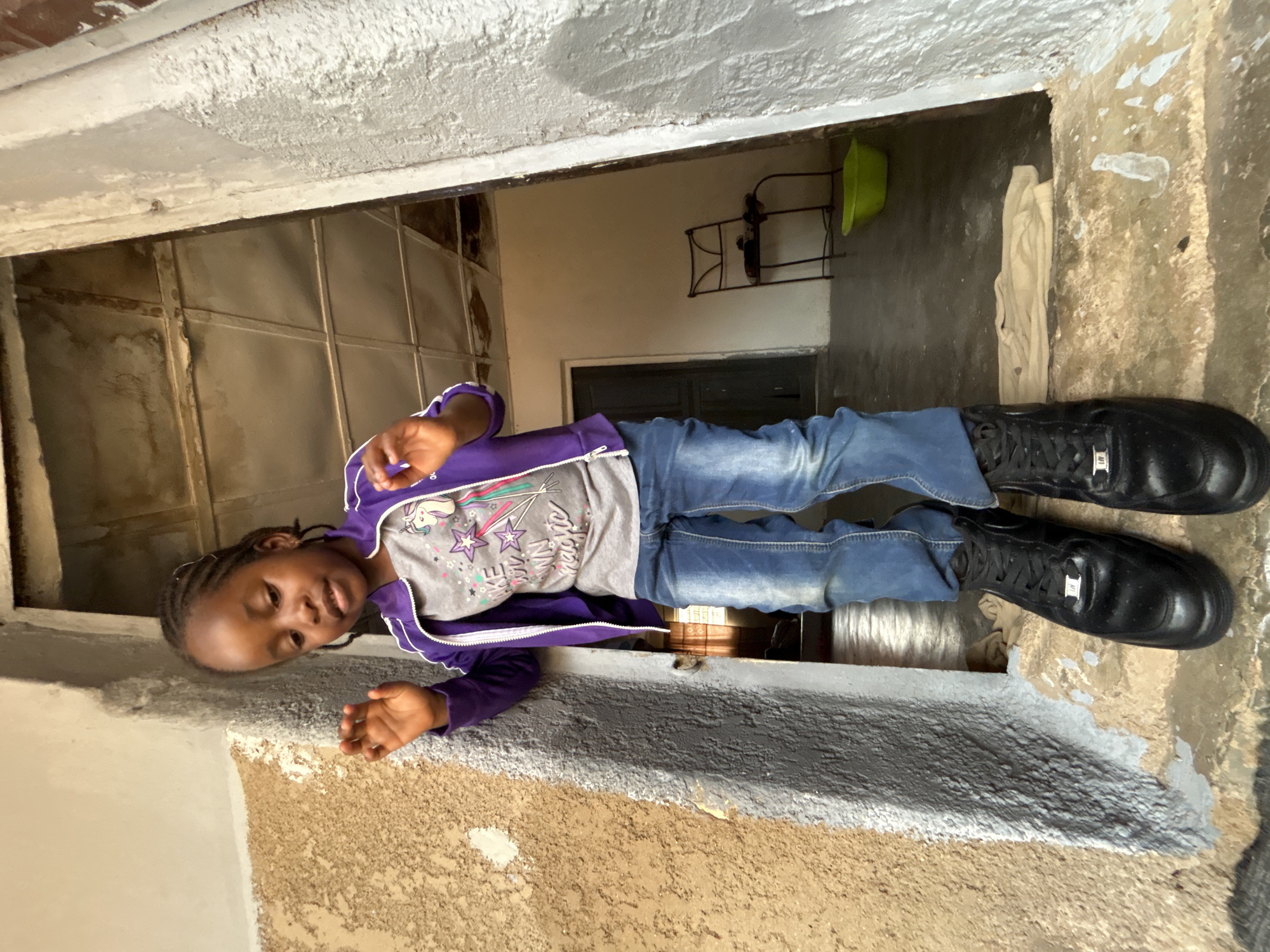 A young, dark-skinned girl wearing a purple hooded sweatshirt over a grey t-shirt with a unicorn and star design and blue jeans, and black sneakers is lying on her side near a partially painted ledge, looking out from a doorway. The room behind the girl is cluttered and includes a metal grid over the opening behind her and objects placed on a metal shelf on the wall. The location is Jos, Nigeria.