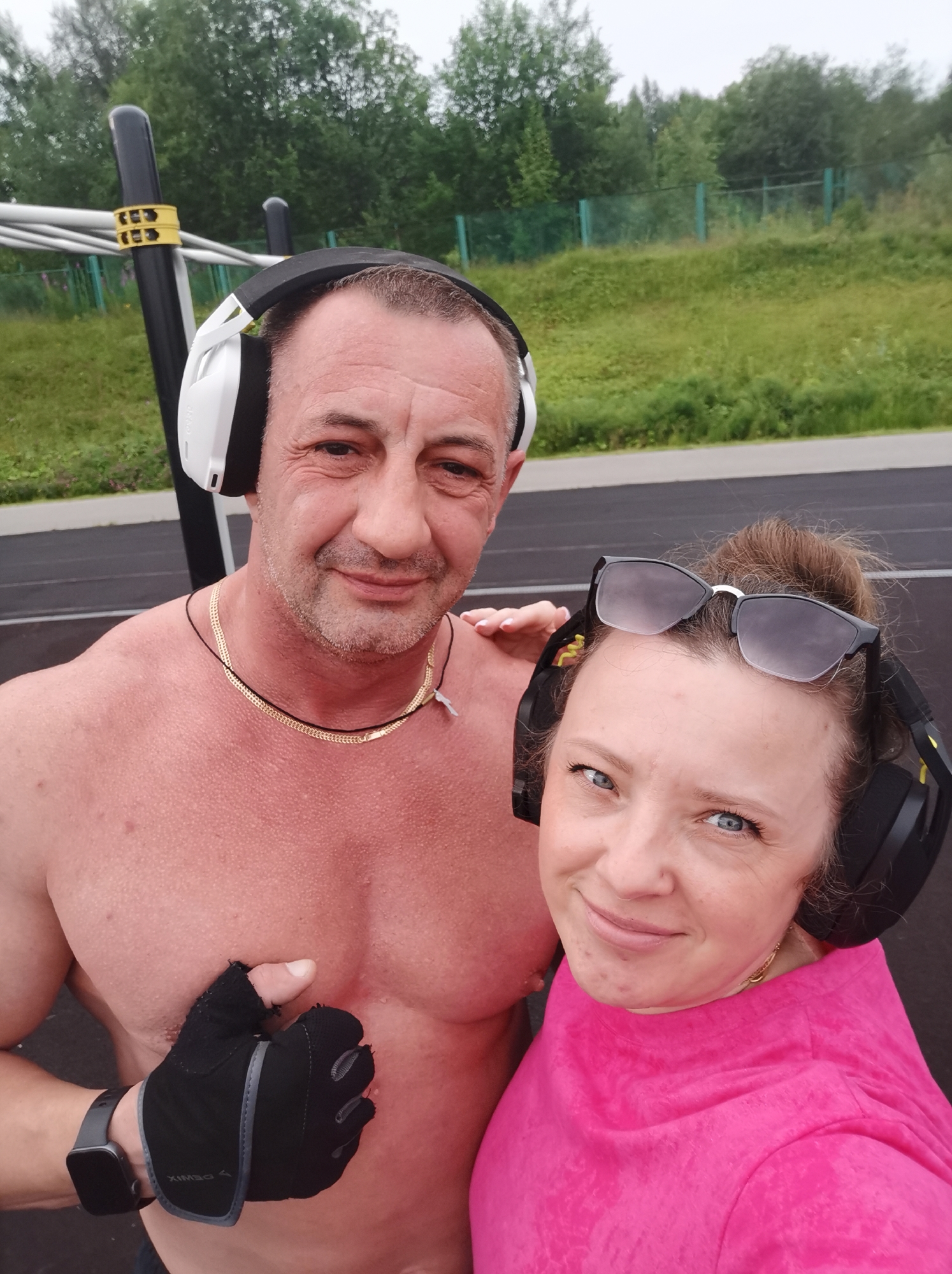 A man and a woman are taking a selfie outdoors. The man, with bare chest and wearing headphones, is on the left, and the woman, in a pink shirt and sunglasses perched on her head, is on the right. They appear to be at an outdoor fitness area with exercise equipment in the background. The environment is a green, grassy area with trees and a fence. The ground is dark asphalt, likely a track or court. The image seems to have been taken in Kizel, Russia, given the context provided.