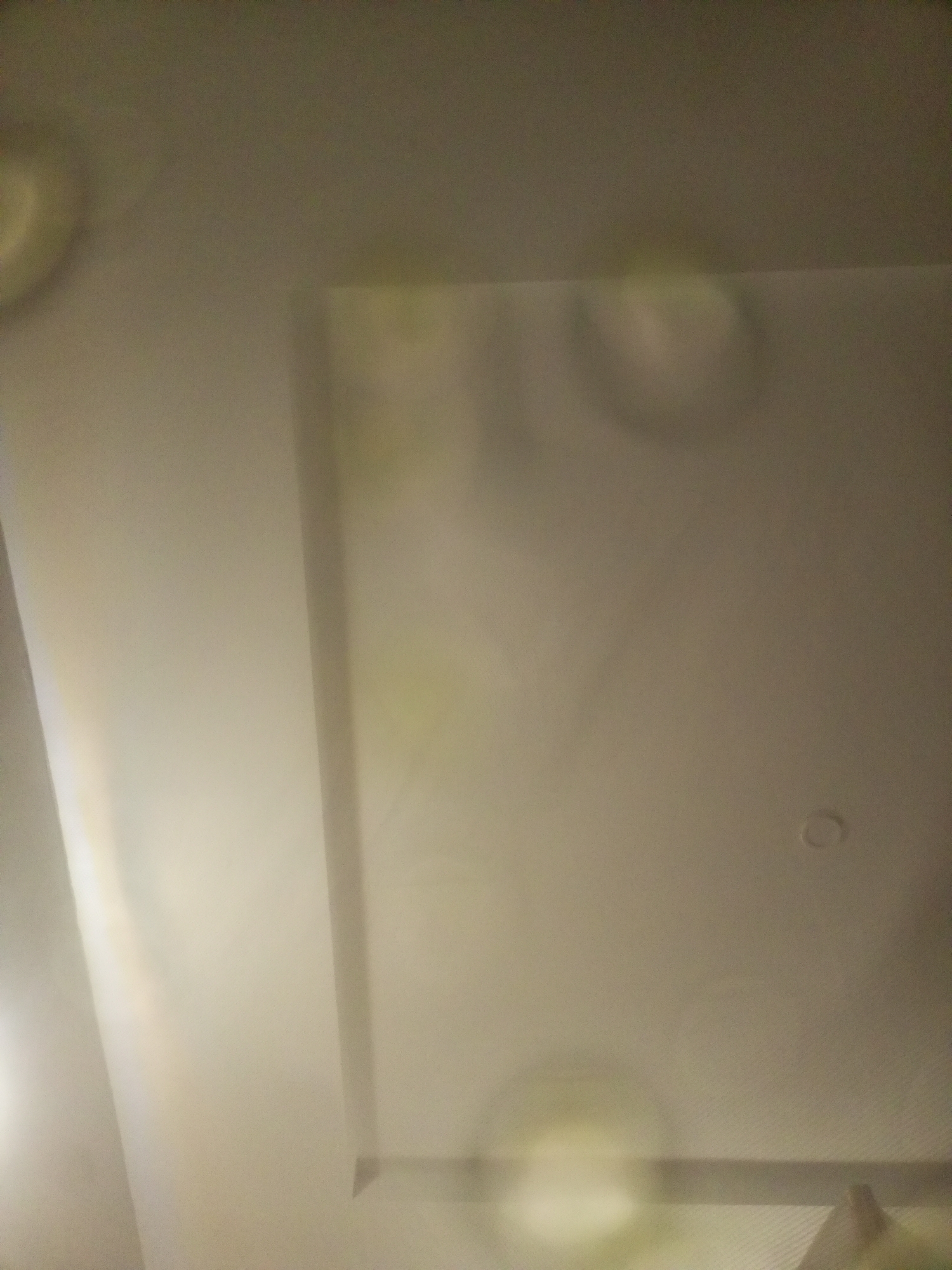 The image shows a ceiling with a recessed area and some subtle lighting effects or reflections. The ceiling appears to be a uniform off-white or light beige color. There is a distinct rectangular recess in the ceiling, with a visible edge. The rest of the ceiling is flat. There are some circular or oval shapes that appear to be light reflections or possibly water stains on the surface of the ceiling, particularly in the recessed area and towards the top left of the image. One small circular fixture, likely a light or smoke detector, is visible on the right side of the flat ceiling. The overall lighting in the scene is dim and diffused. The image is taken from a low angle, looking upwards towards the ceiling. The location is Unknown, Unknown.