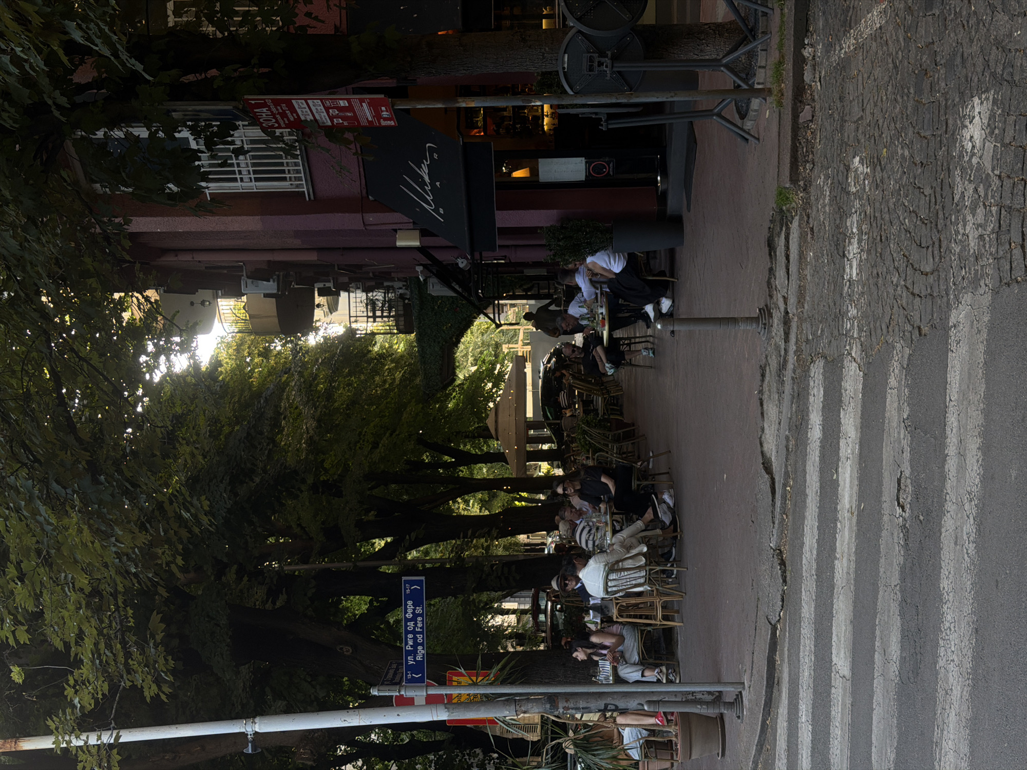 The image depicts an outdoor café on a street in Belgrade, Serbia. Tables and chairs are arranged along the sidewalk, and several people are seated, some engaged in conversation. Trees line the street, casting shadows and adding to the atmosphere. A street sign indicates "ul. Riga od Fere St.", identifying the location as Rige od Fere Street. A crosswalk is visible on the right side of the image. The building adjacent to the café is a burgundy-colored building with an awning marked "Mikan".
