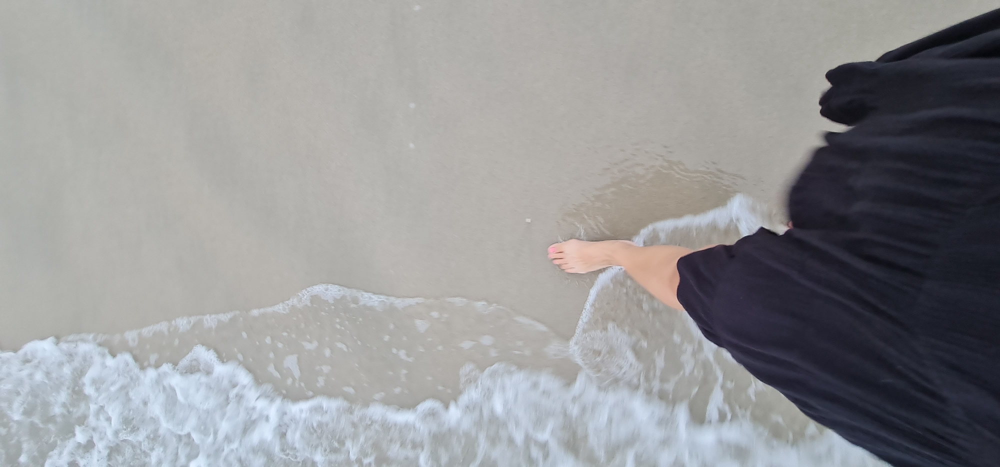 A top-down, slightly tilted perspective captures a scene on a beach in Đà Nẵng, Vietnam. White sand is visible, with the ocean lapping the shore, creating white foamy waves. A fair-skinned foot with painted pink toenails is partially submerged in the water. The right side of the image is dominated by a flowing, dark fabric, possibly a dress or skirt.